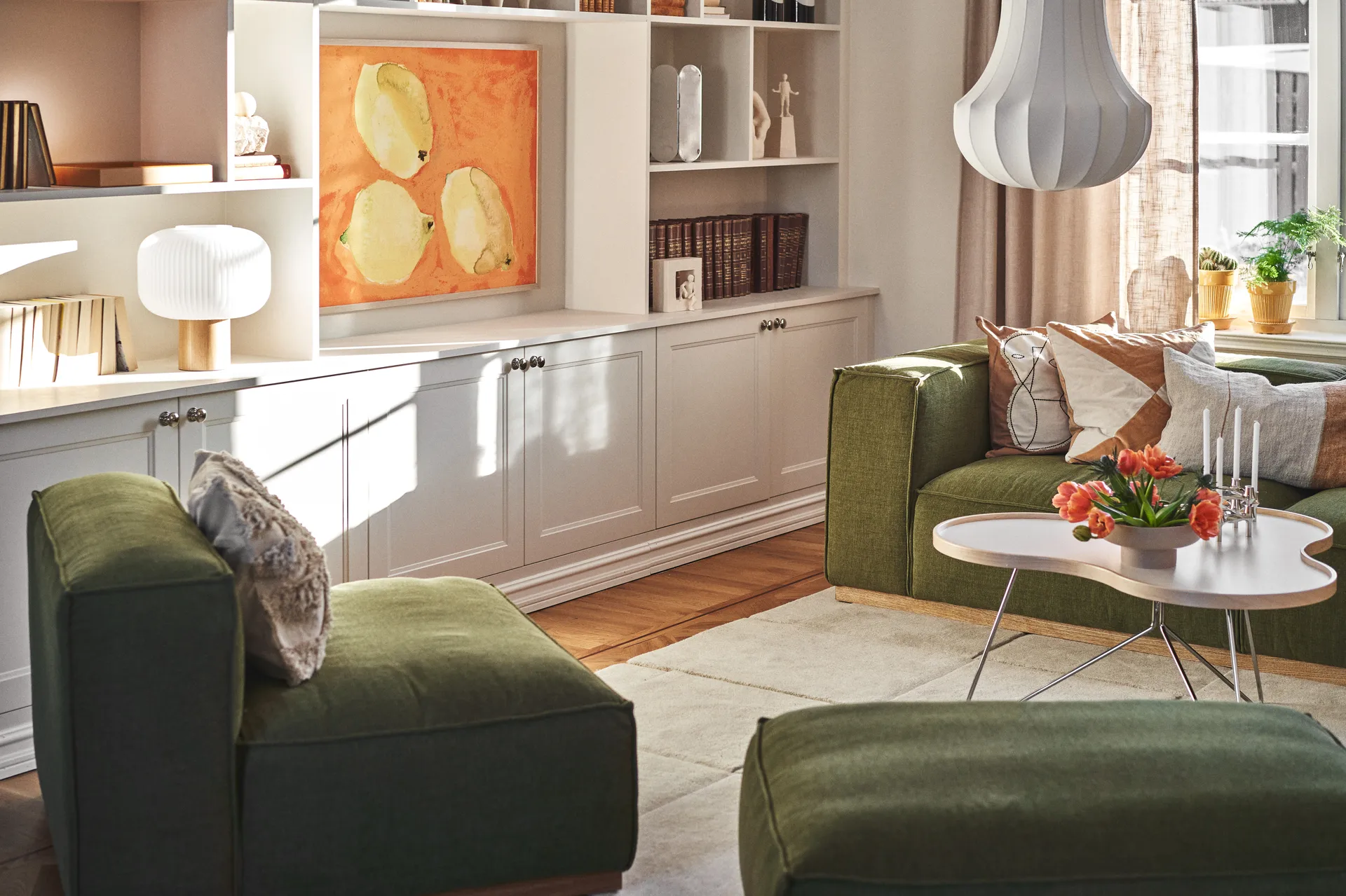 Aurinkoinen olohuone vihreillä sohvilla, valkoinen sohvapöytä tulppaaneilla ja kynttilöillä, valkoiset hyllyt taiteella.