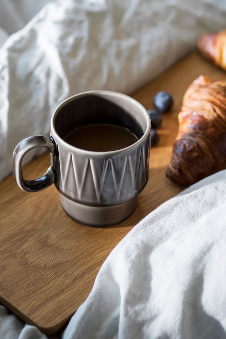 Harmaa Coffee & More -espressokuppi seisoo puulevyllä sängyssä, ja sen vieressä on croissant.
