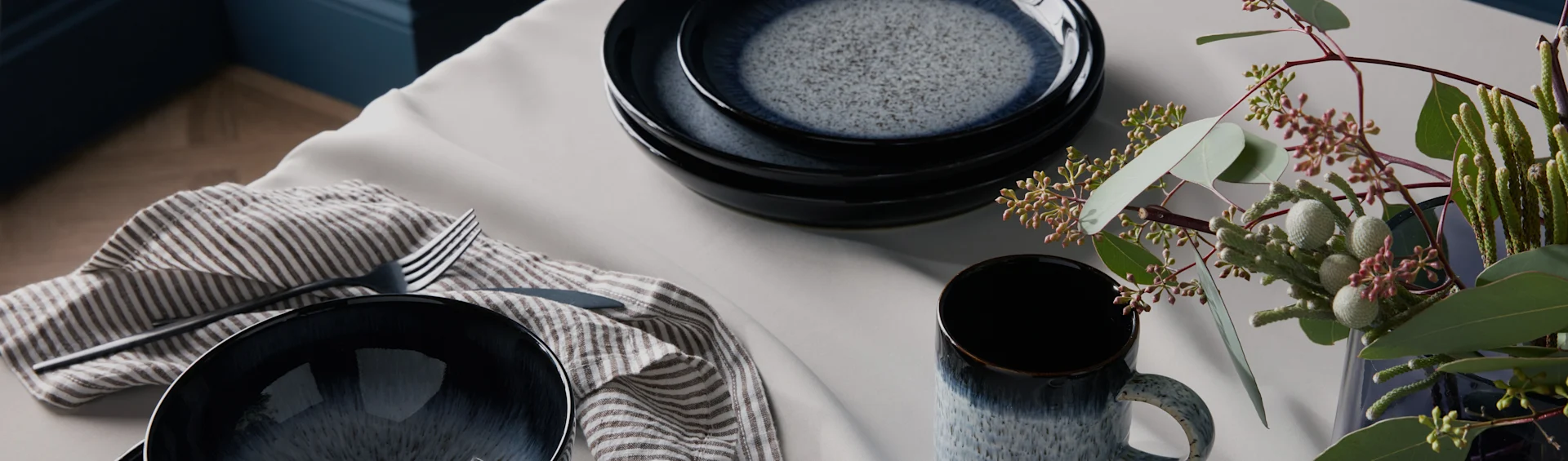 Pilkkuneliöinen tummansininen keramiikka-astiasto, raidallinen lautasliina, ruokailuvälineet ja vihreitä lehtiä vaalealla pöydällä.