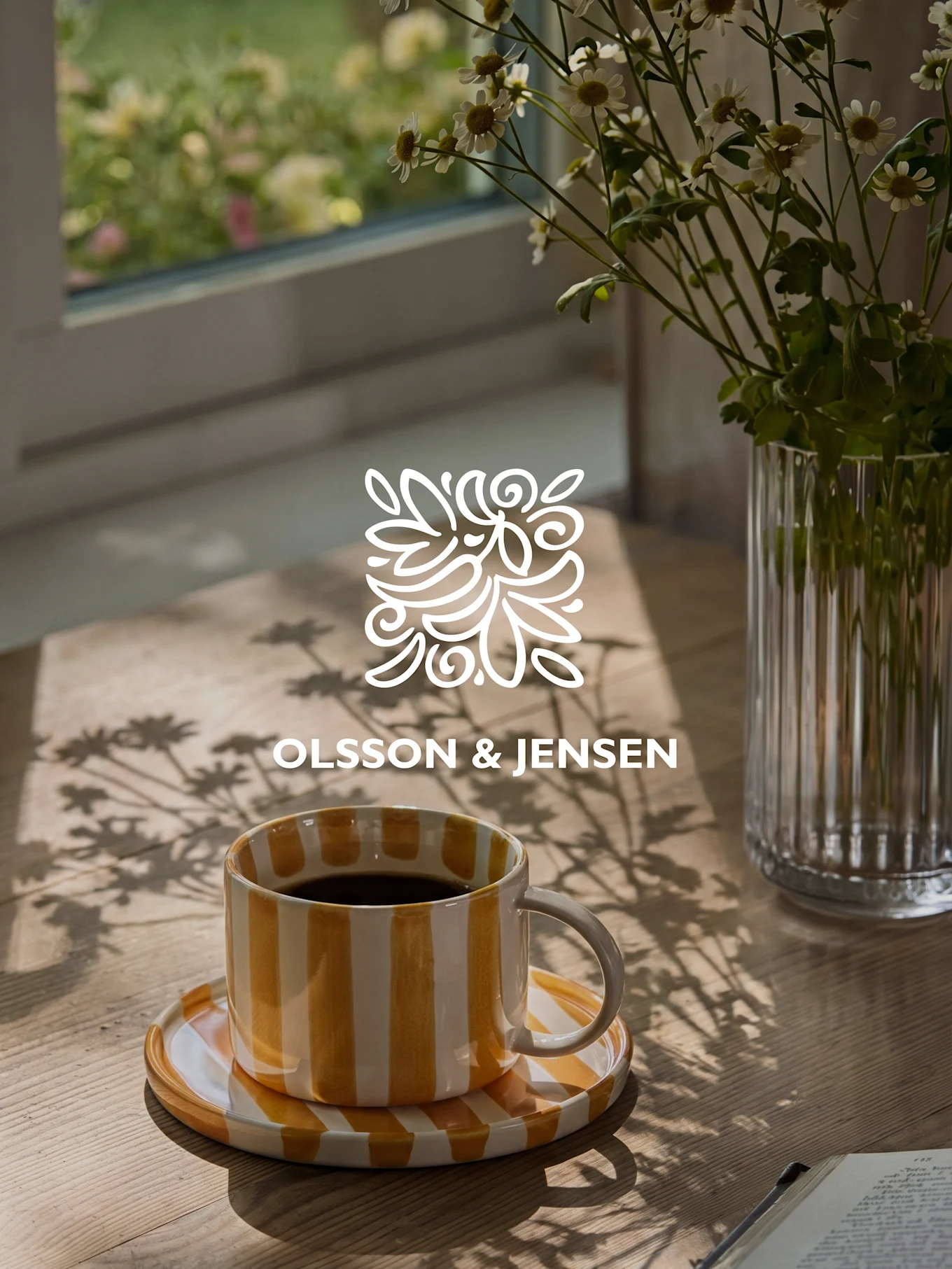 Kelta-valkoraidallinen kahvimuki puupöydällä valkoisten kukkien ja auringonvalon varjojen kanssa, Olsson & Jensen -logo.