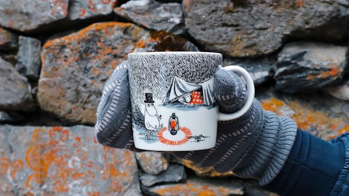 @moominbyingri pitää Schlaf gut Moomin -kuppia kivenseinän edessä käsine kädessä.