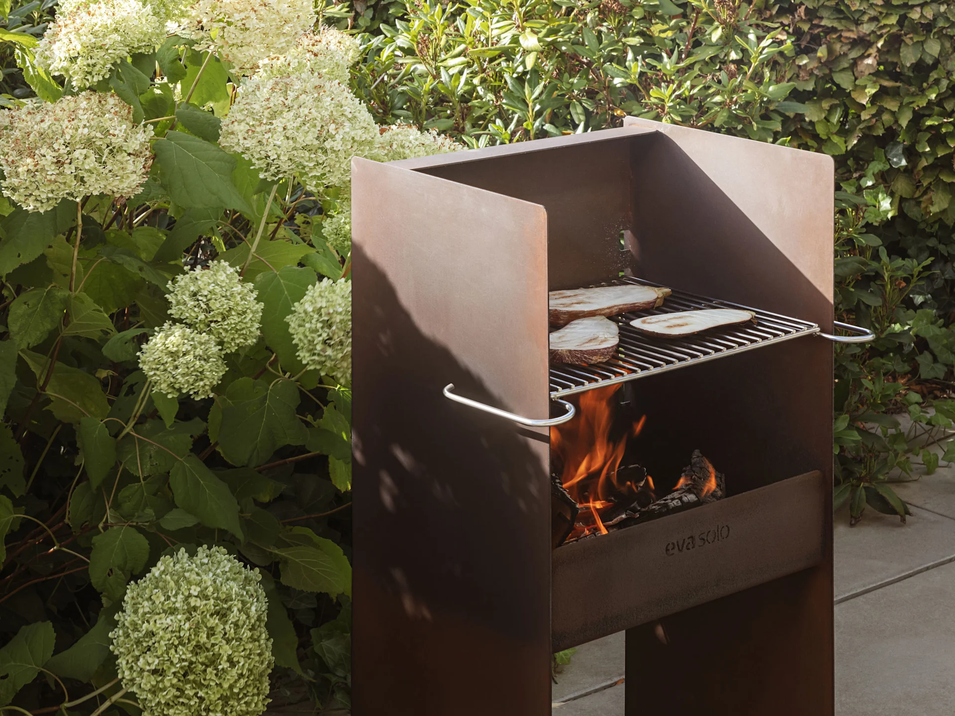 Rustiikkinen metallinen ulkogrilli, jossa on liekkejä ja munakoisoviipaleita paistumassa ritilällä.