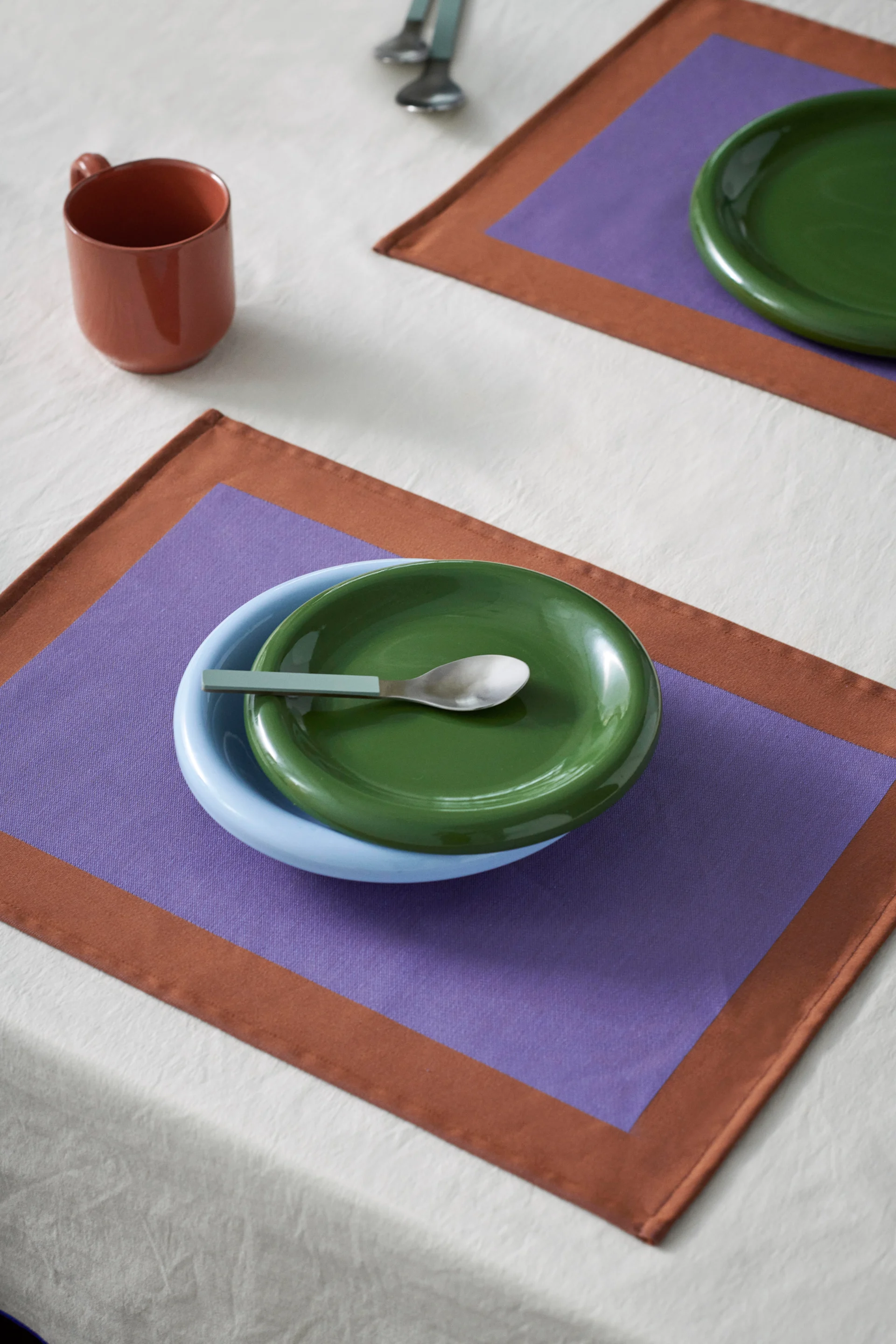 Pöytäkattaus sinisellä kulholla, vihreällä lautasella ja lusikalla purppuranruskealla tableteella. Ruskea muki valkoisella liinalla.