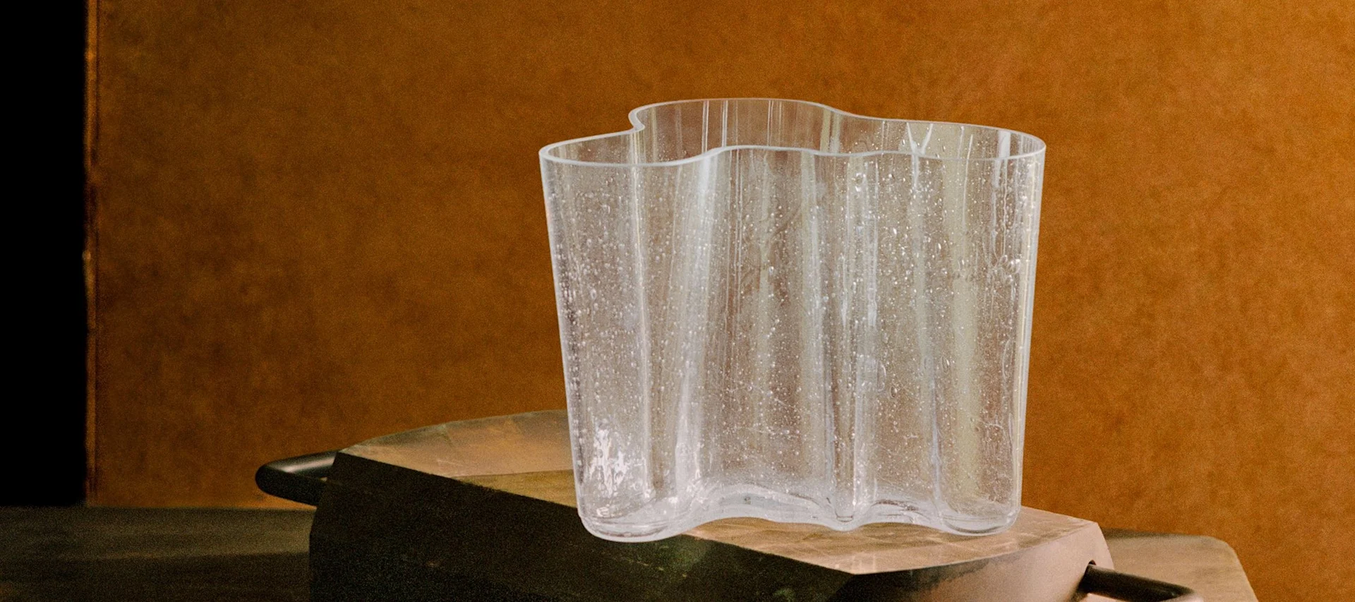 Kirkas, aaltoileva lasimaljakko, jossa on teksturoituja läikkiä, rustiikkisella tummalla pinnalla lämpimänruskeaa taustaa vasten.