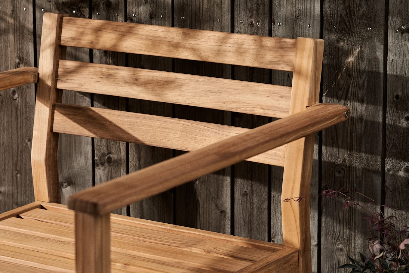 Rimoitettu puutuoli käsinojilla tummaa puuaitaa vasten, peitetty lehtien varjoilla.