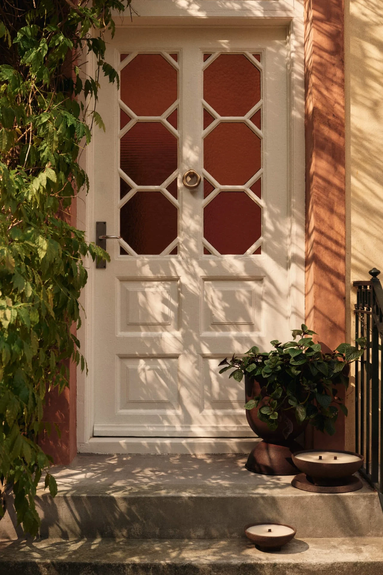 Valkoinen etuovi kahdeksankulmaisilla lasipaneeleilla, vihreitä köynnöksiä vasemmalla ja istutusastia kahdella kynttilällä portailla.