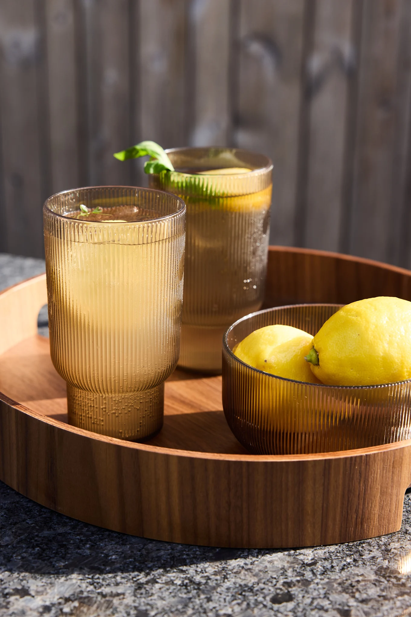 Kaksi uurrettua meripihkanväristä lasia limonadilla ja kulho sitruunoita puisella tarjottimella ulkona.