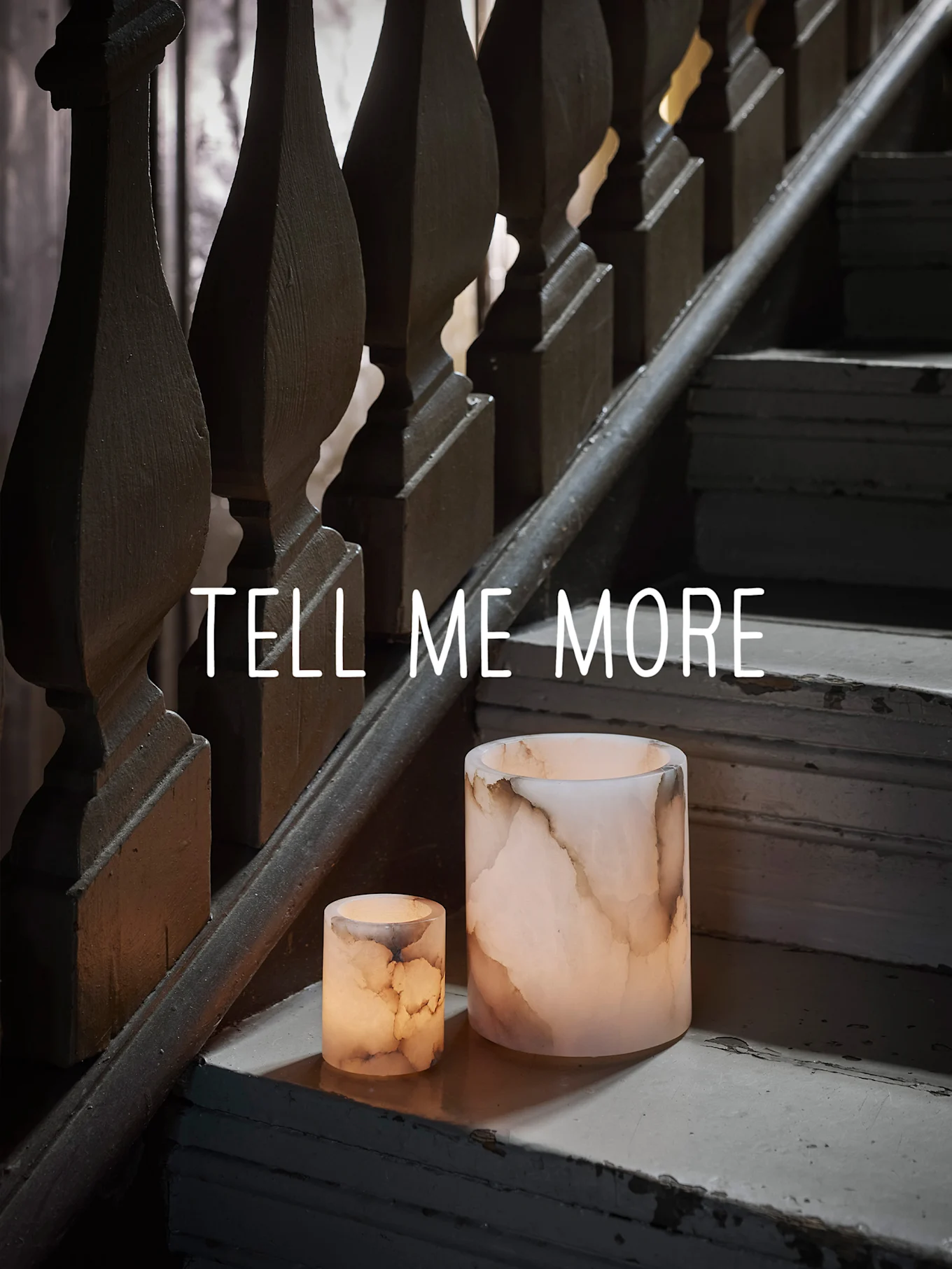 Kaksi hehkuvaa onyksimäistä kynttilänjalkaa kuluneilla puuportailla, tekstillä "TELL ME MORE".