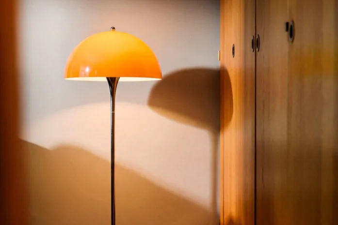 Retrohenkinen lattiavalaisin oranssilla kupuvarjostimella ja kromijalalla valaisee huonetta, jossa on kuvioitu matto ja puinen vaatekaappi.