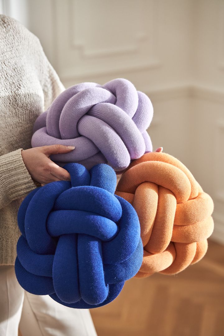Design House Stockholm merkin sinisiä, violetteja ja aprikoosinvärisiä Knot-tyynyjä naisen sylissä.