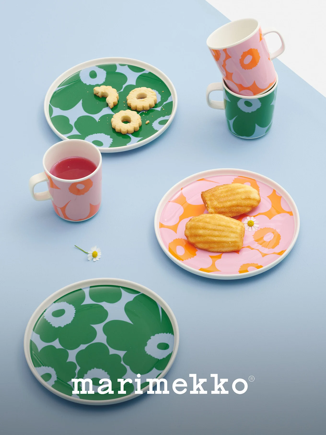 Marimekko-lautaset ja -mukit rohkeilla kukkakuvioilla vihreässä, vaaleanpunaisessa ja oranssissa, sisältäen keksejä, madeleine-leivoksia ja juoman.