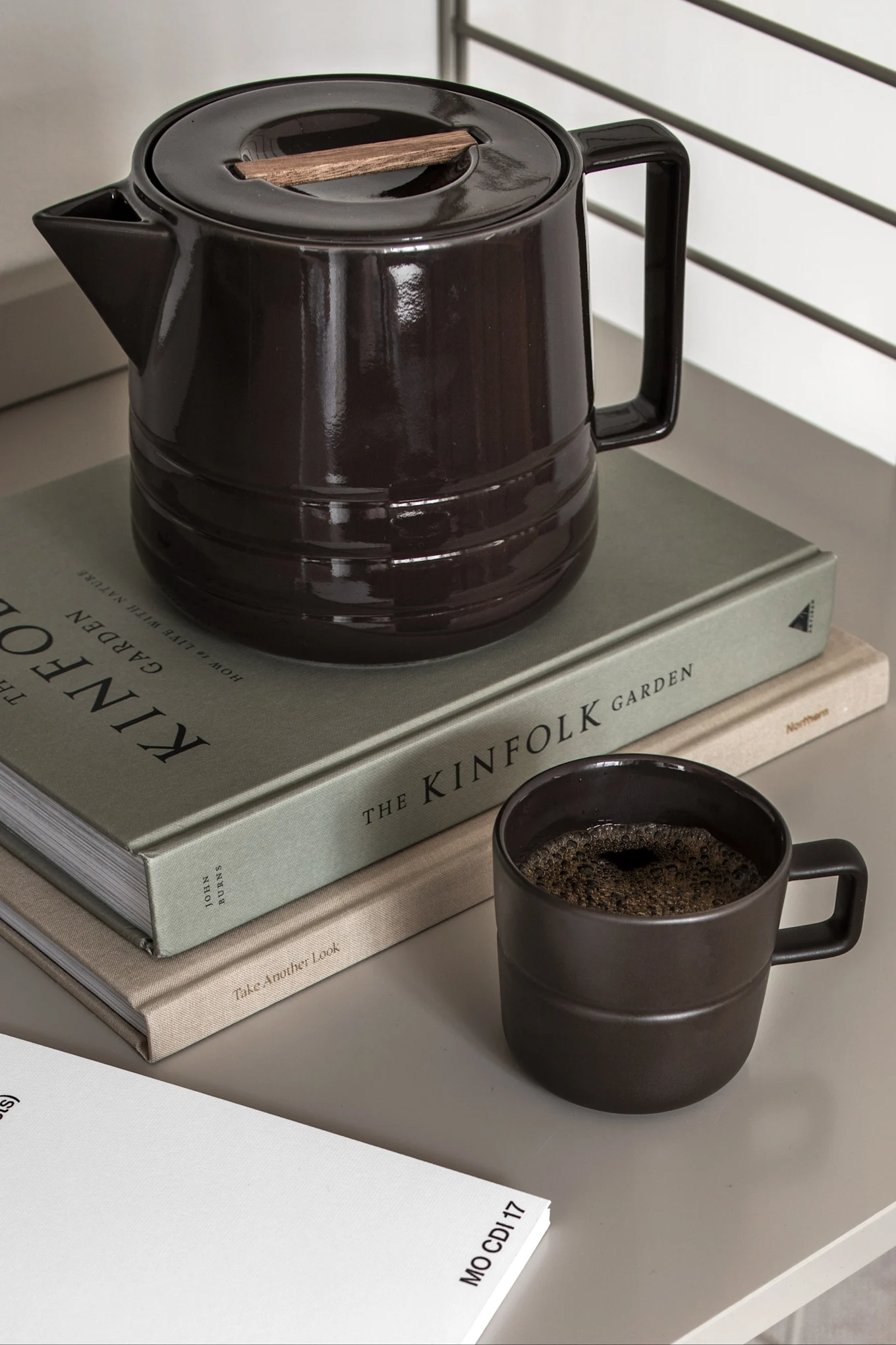 Tummanruskea keraaminen kahvipannu puisella kannella, yhteensopiva muki kahvin kanssa ja kaksi kirjaa.