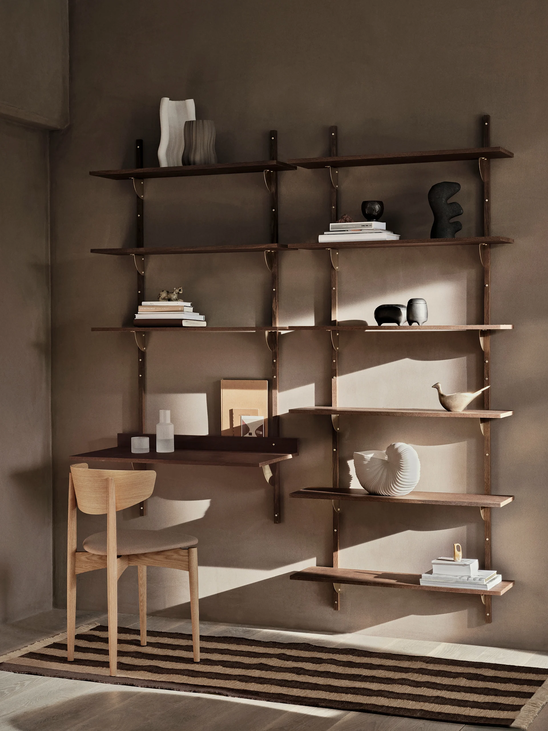 Moderni tumman puinen seinähyllykkö työpöydällä ja tuolilla, koristeltu kirjoilla ja koriste-esineillä, on lämpimän ruskeaa seinää vasten raidallisen maton kera.