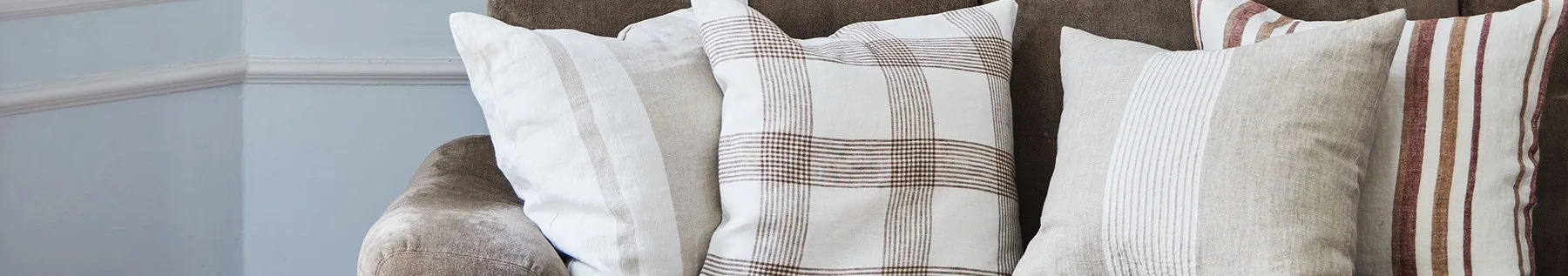 Lähikuva kolmesta tyynystä ruskealla sohvalla, joissa valkoisia, beigenvärisiä ja ruudullisia kuvioita ruskeilla ja punaisilla raidoilla.