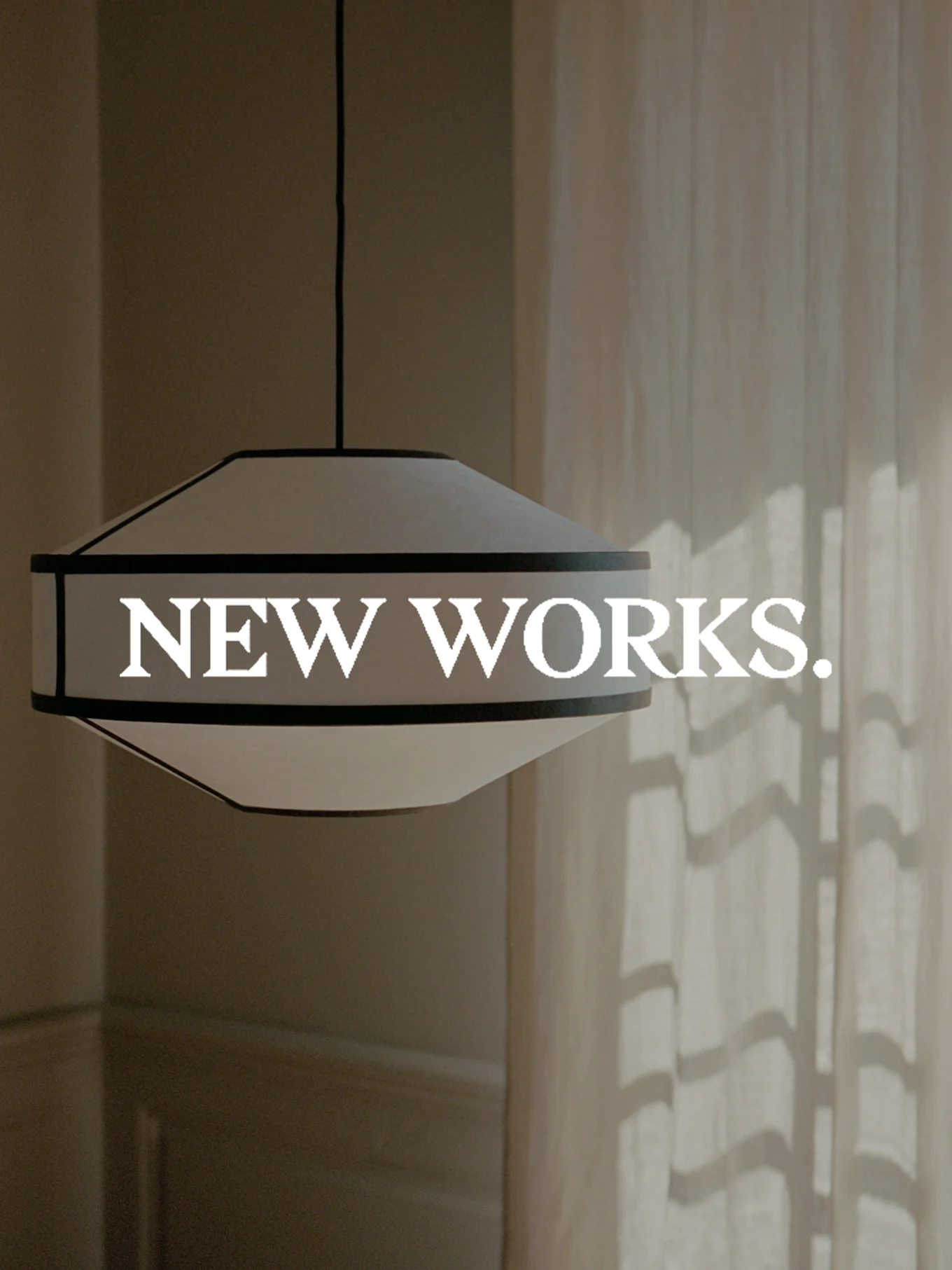 Leveä, valkoinen ja tummareunainen riippuvalaisin roikkuu, ja siinä on teksti 'NEW WORKS.' päällä. Auringonvalo heittää varjoja verhoon.