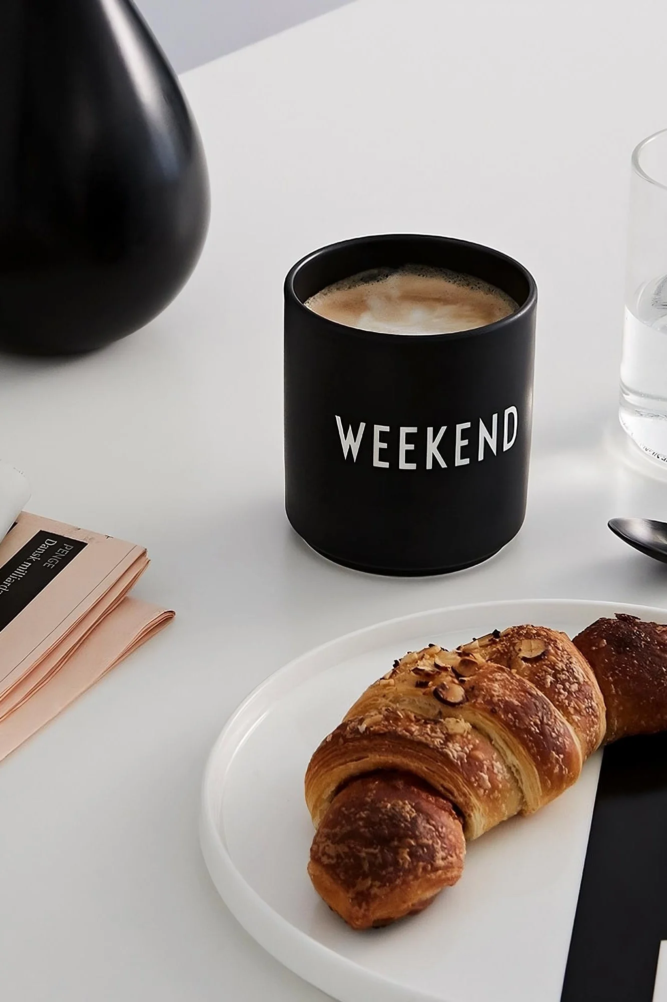 Kahvimuki: Design Lettersin musta kahvimuki, jossa on teksti ”Weekend”, seisoo pöydällä, ja sen edessä on lautasellinen croissanteja.