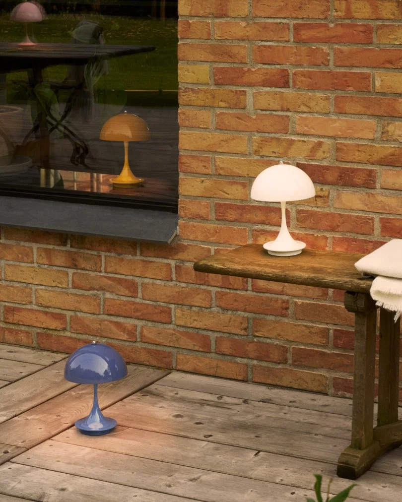 Kolme sienimäistä lamppua valkoisena, sinisenä ja keltaisena puisella patiolla tiiliseinän ja ikkunan vieressä.
