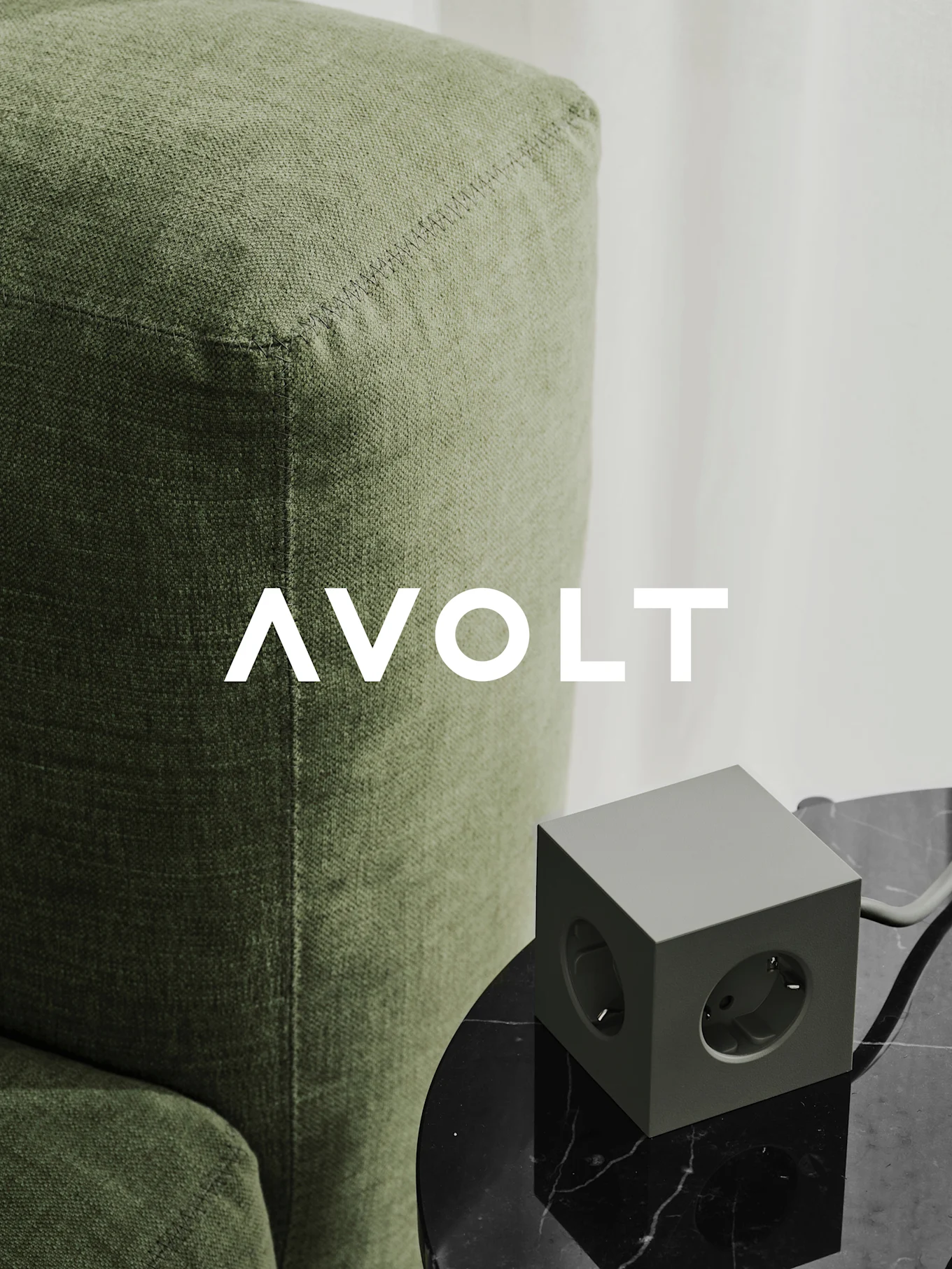 Vihreä kuutionmuotoinen jatkojohto kolmella pistorasialla mustalla marmoripöydällä vihreän sohvan vieressä. AVOLT-logo.
