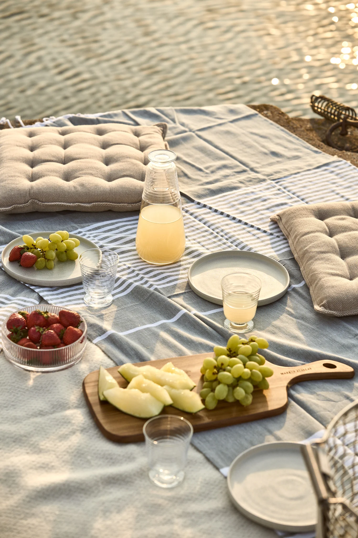Piknik veden äärellä raidallisen peiton, tyynyjen, mehun, mansikoiden, viinirypäleiden ja meloniviipaleiden kera.