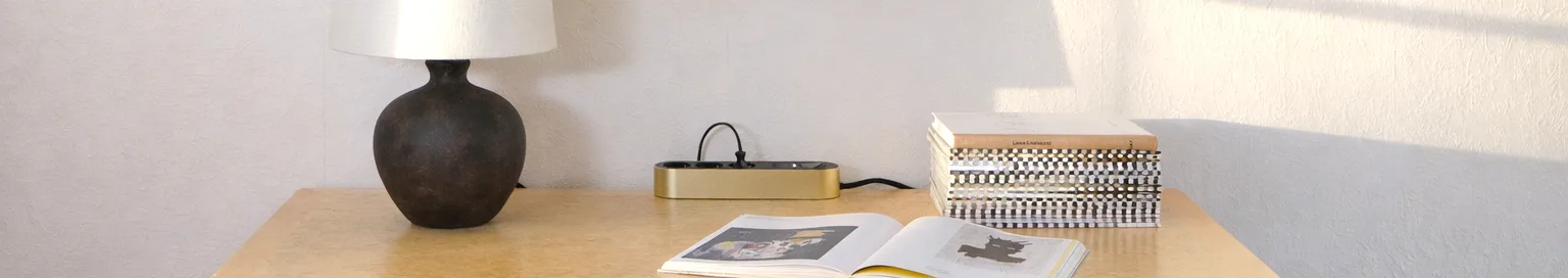 Kultainen jatkojohto kolmella mustalla pistorasialla ja virtapainikkeella vaalealla pinnalla.