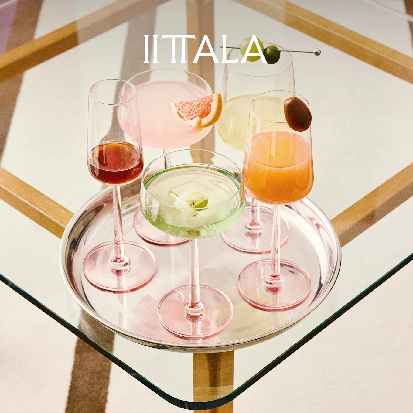 IITTALA-logo hopeatarjottimen yläpuolella, jossa viisi värikästä cocktailia vaaleanpunaisilla varsilla olevissa laseissa lasipöydällä.