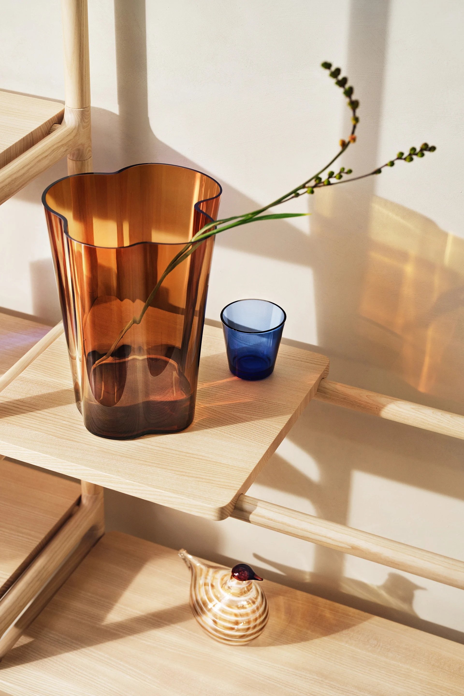 Meripihkanvärinen lasimaljakko, jossa on aaltoileva reuna ja oksa, sekä sininen lasiesine ja raidallinen lasilintu vaalealla puuhyllyllä.
