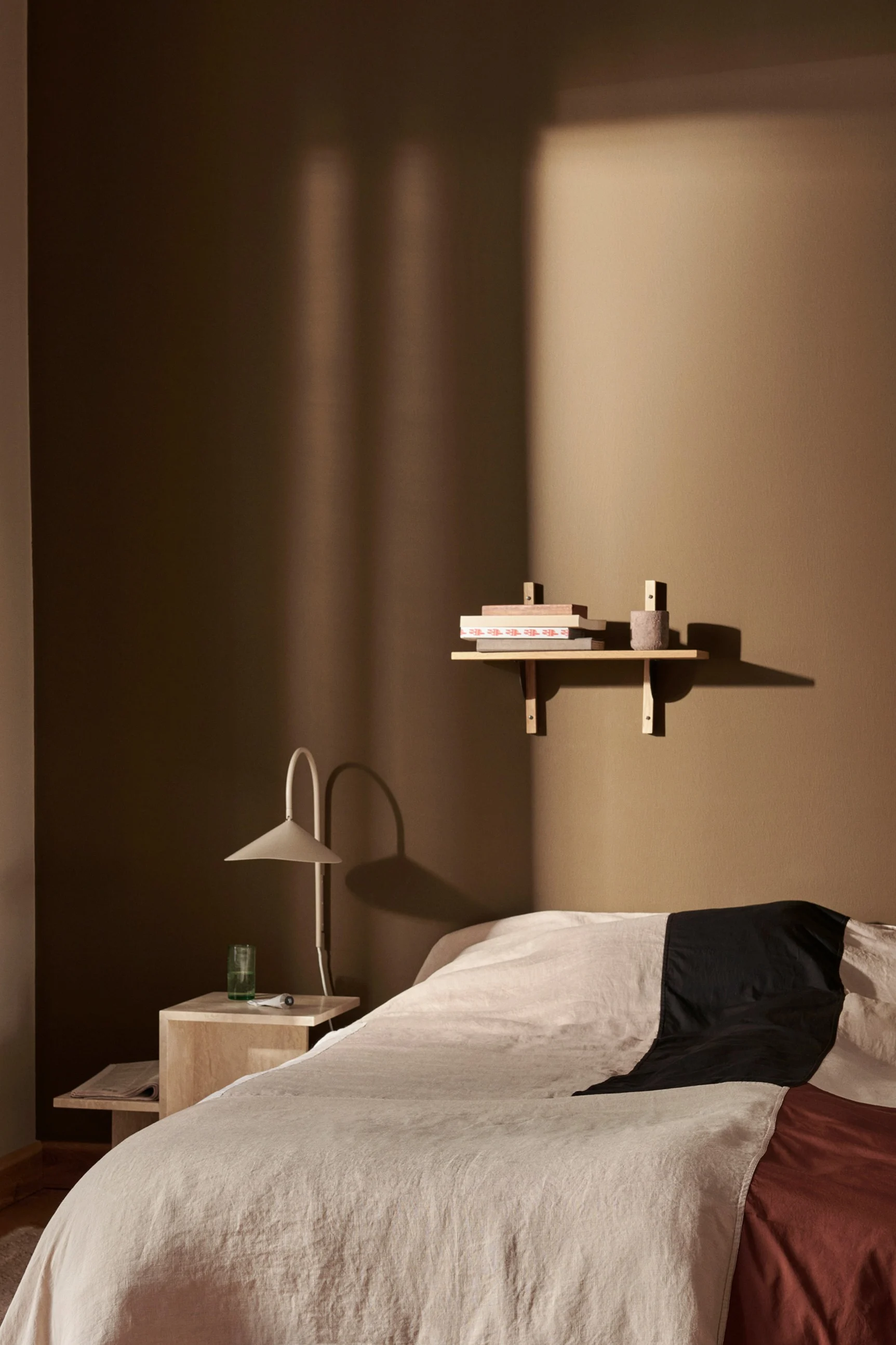 Makuuhuone, jossa sänky beigeillä, mustilla ja ruosteenruskeilla vuodevaatteilla, yöpöytä lampun kanssa sekä hylly kirjoineen lämpimänruskealla seinällä. Koristele makuuhuoneen seinä seinävalaisimella ja hyllyllä.