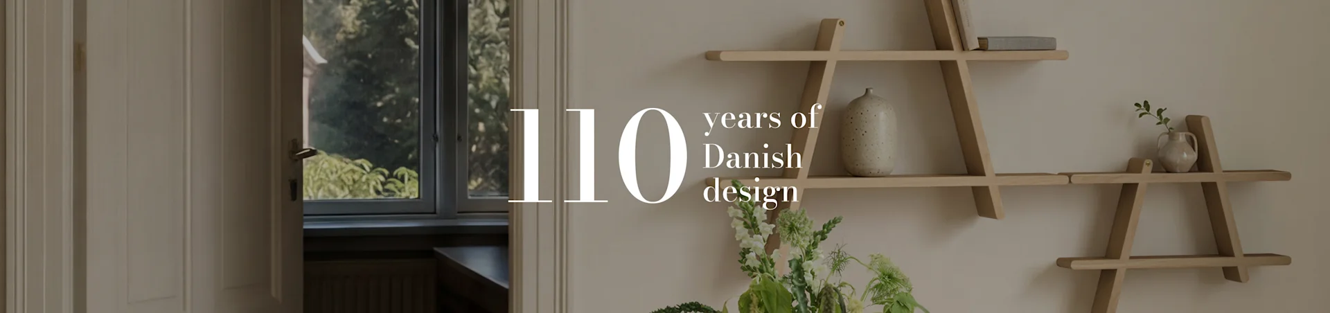 Teksti '110 years of Danish design' huoneen sisäkuvan päällä, jossa on puinen geometrinen hylly, maljakko ja kukkia.