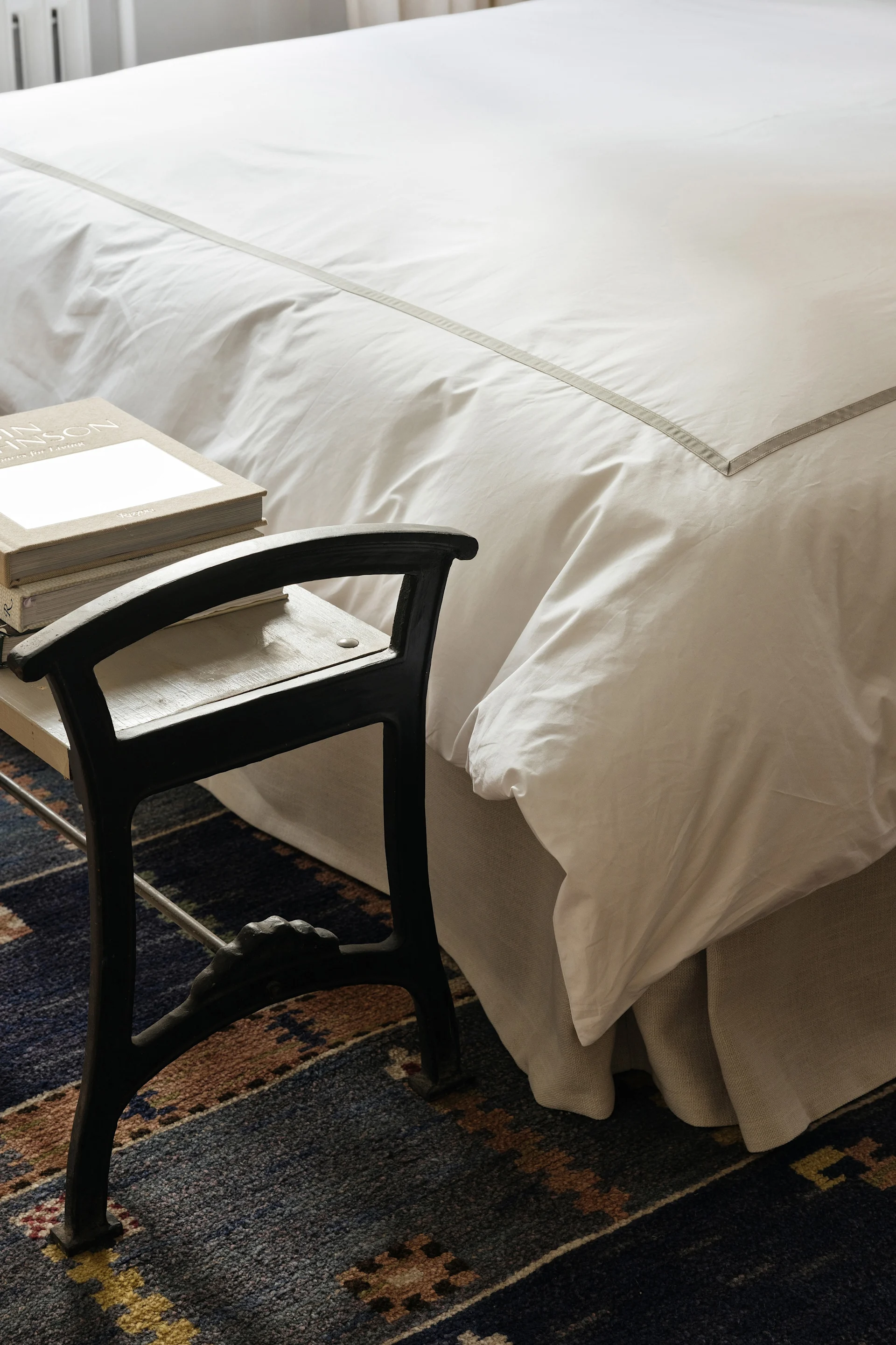 Sänky, jossa on valkoiset vuodevaatteet ja harmaa raita, vieressä tumma jakkara, jonka päällä on kirjoja, kuviollisella sinisellä matolla.