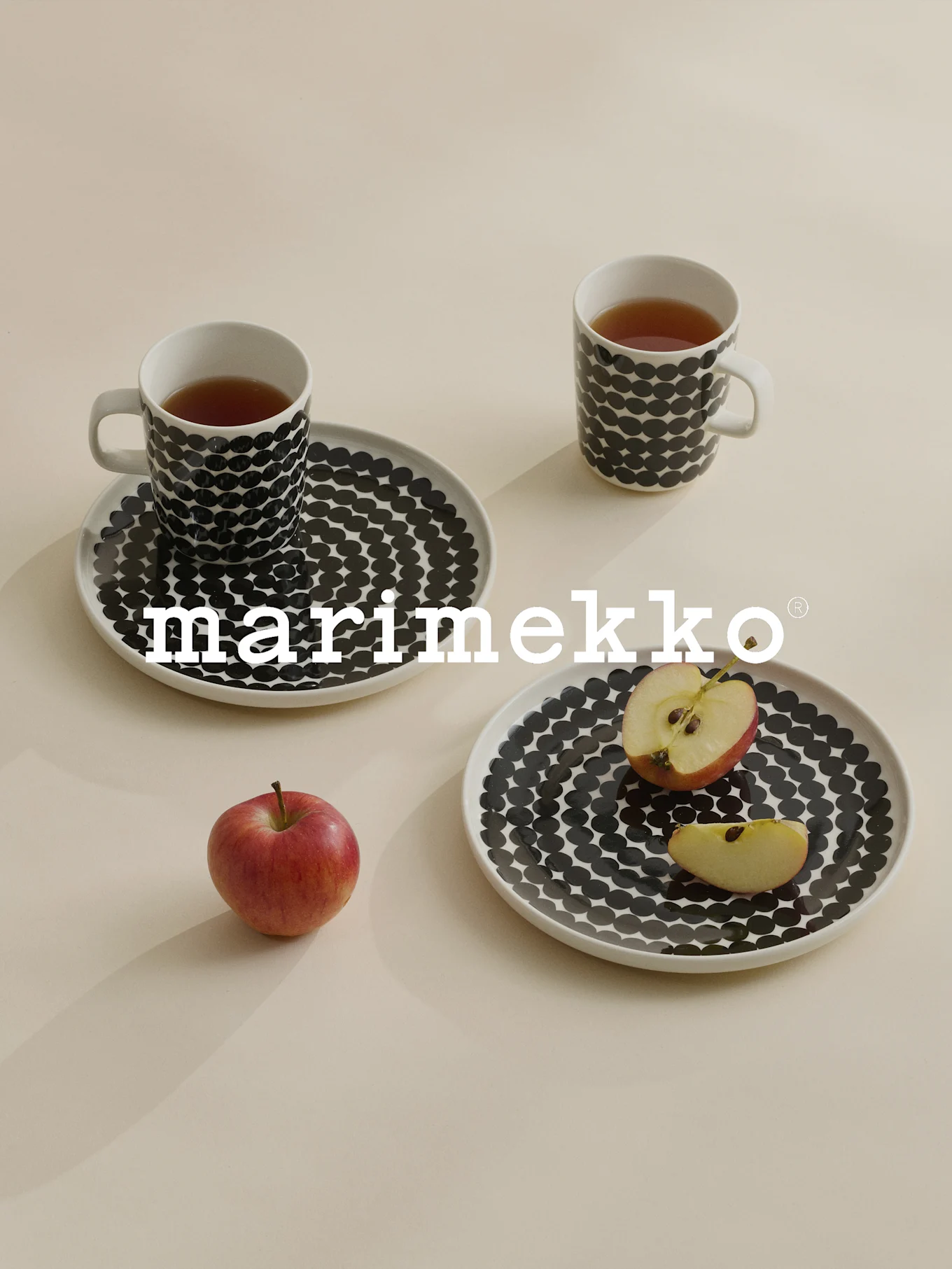 Kaksi mukia ja lautasta mustilla pilkuilla, teetä, punainen omena, Marimekko-logo.
