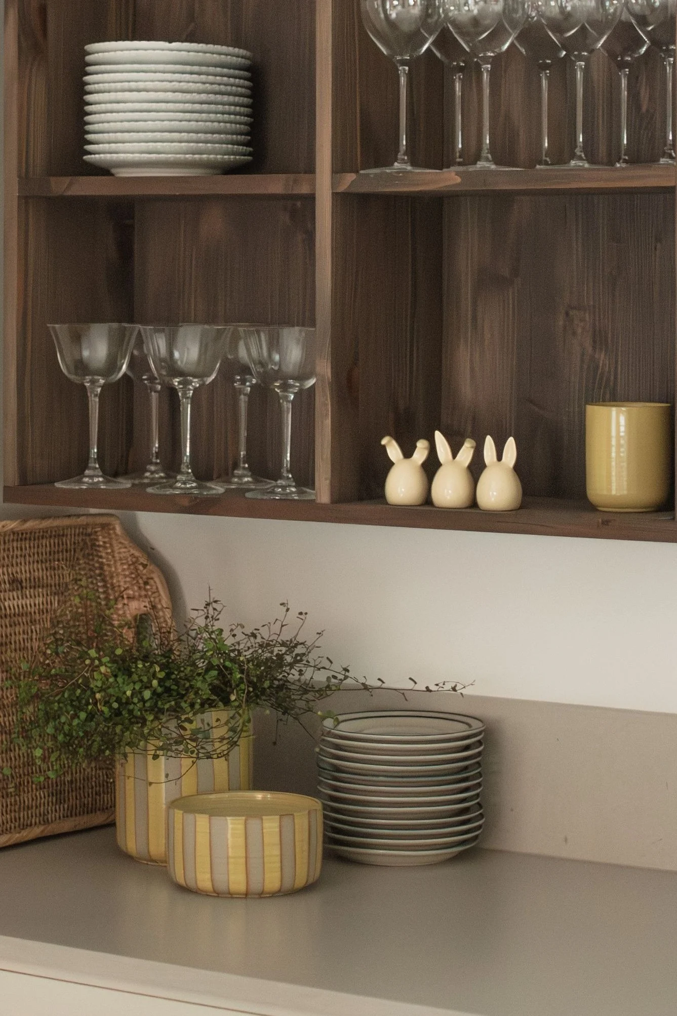 Puuhyllyillä pinottuja valkoisia lautasia, erilaisia laseja, kolme keraamista pupua ja keltainen muki. Tiskillä on punottu kori, kasveja raidallisissa ruukuissa ja lisää lautasia.