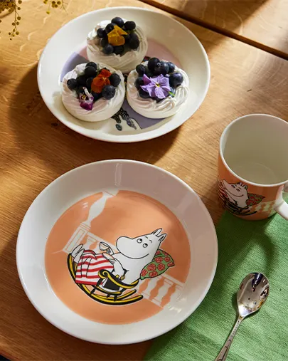 Muumi-lautanen, muki ja lusikka lautasen vieressä, jossa on marenkeja mustikoilla ja kukilla puupöydällä.