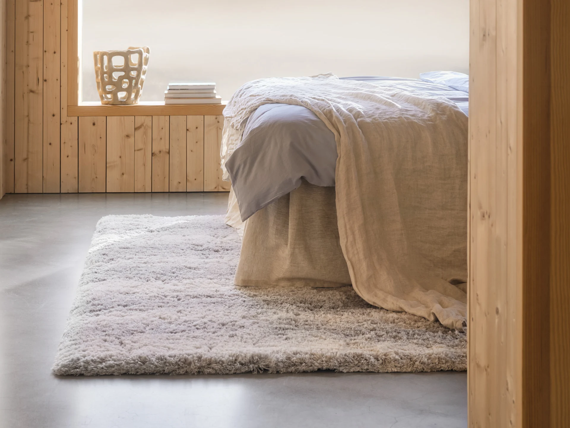 Viihtyisä makuuhuone, jossa sänky, vaaleansiniset lakanat, beige viltti, pörröinen matto, puupaneloidut seinät ja koristeellinen maljakko.