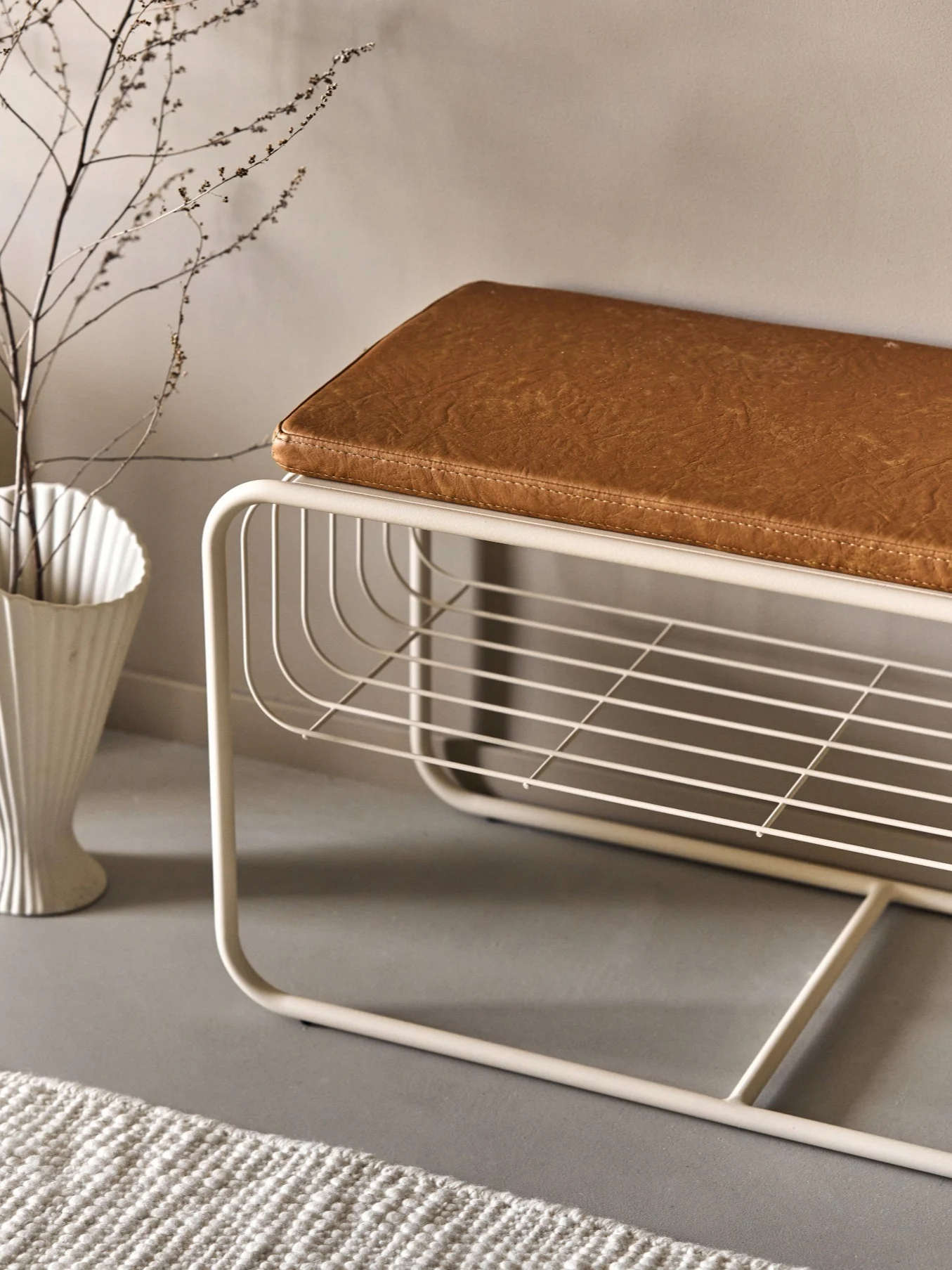 Moderni beige metallipenkki, jossa ruskea tekonahkainen tyyny ja avoin säleikköhylly. Valkoinen maljakko oksineen ja matto ovat lähellä.