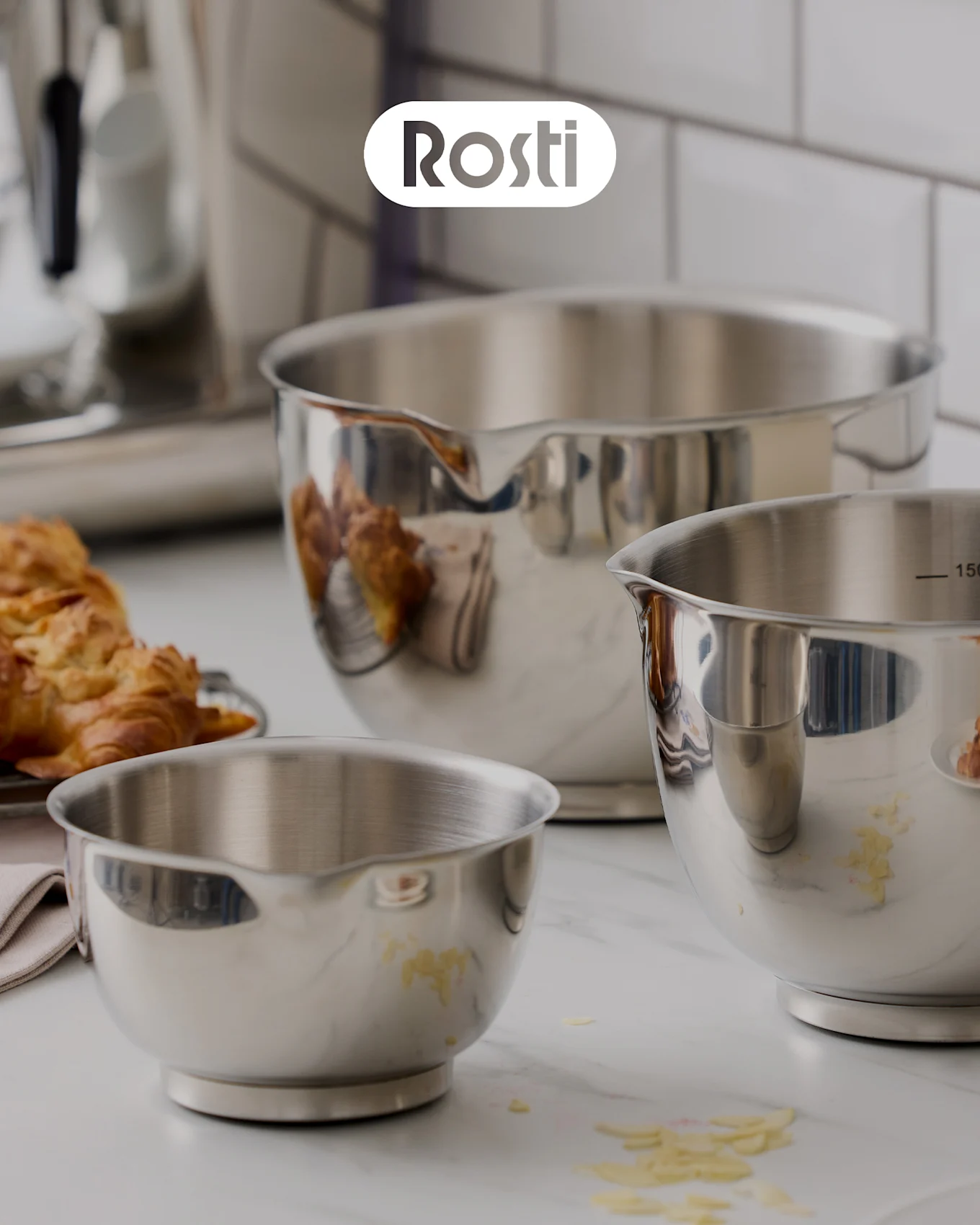 Kolme Rosti-kulhoa tiskillä: beige kulho, jonka päällä on sininen, molemmissa kannet, sekä pieni keltainen kulho täynnä kuivattuja aprikooseja.