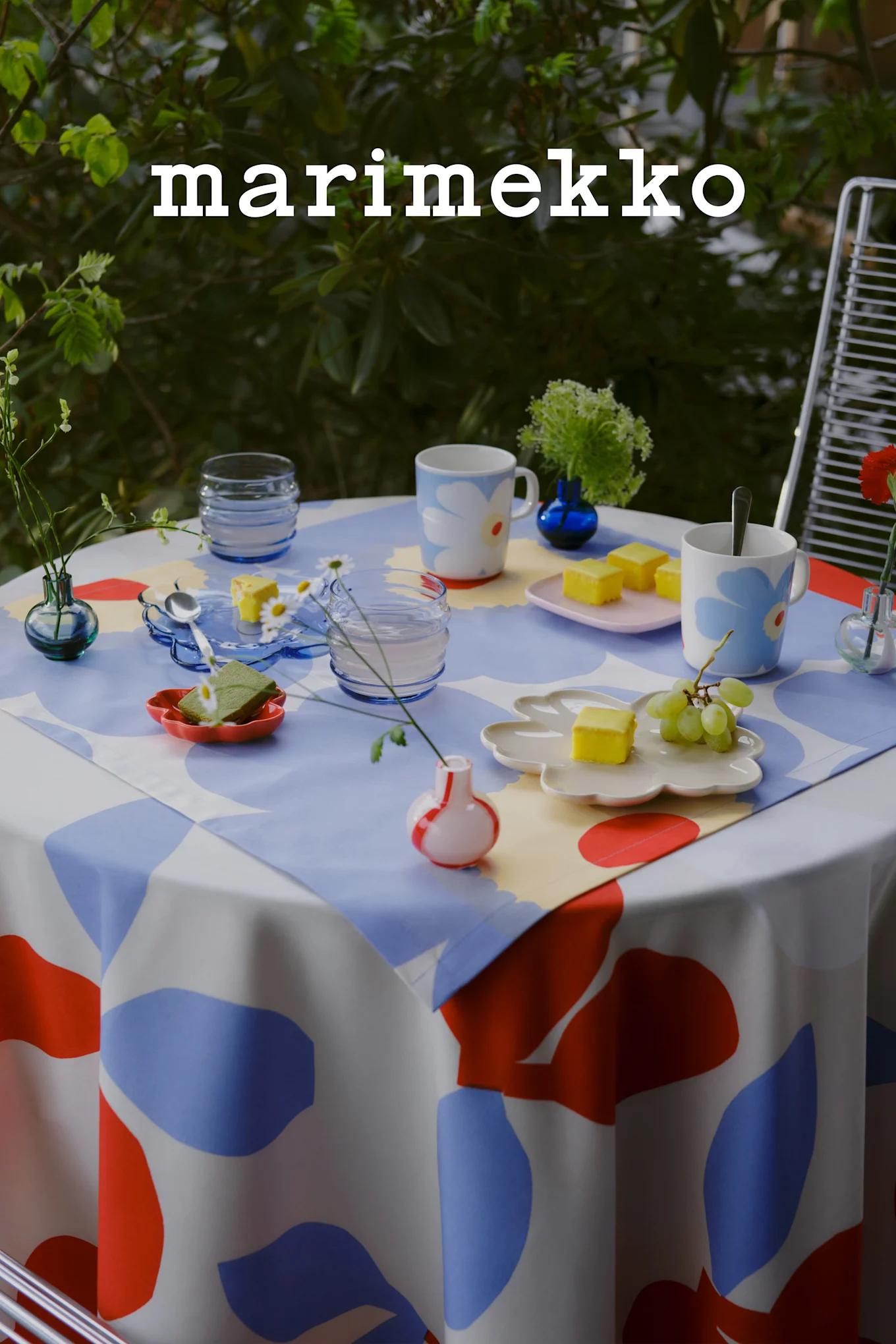 Marimekko-logo. Ulkopöytä sinivalko-punaisella abstraktilla liinalla, kukkamukeilla, kakuilla, rypäleillä, pienillä maljakoilla.
