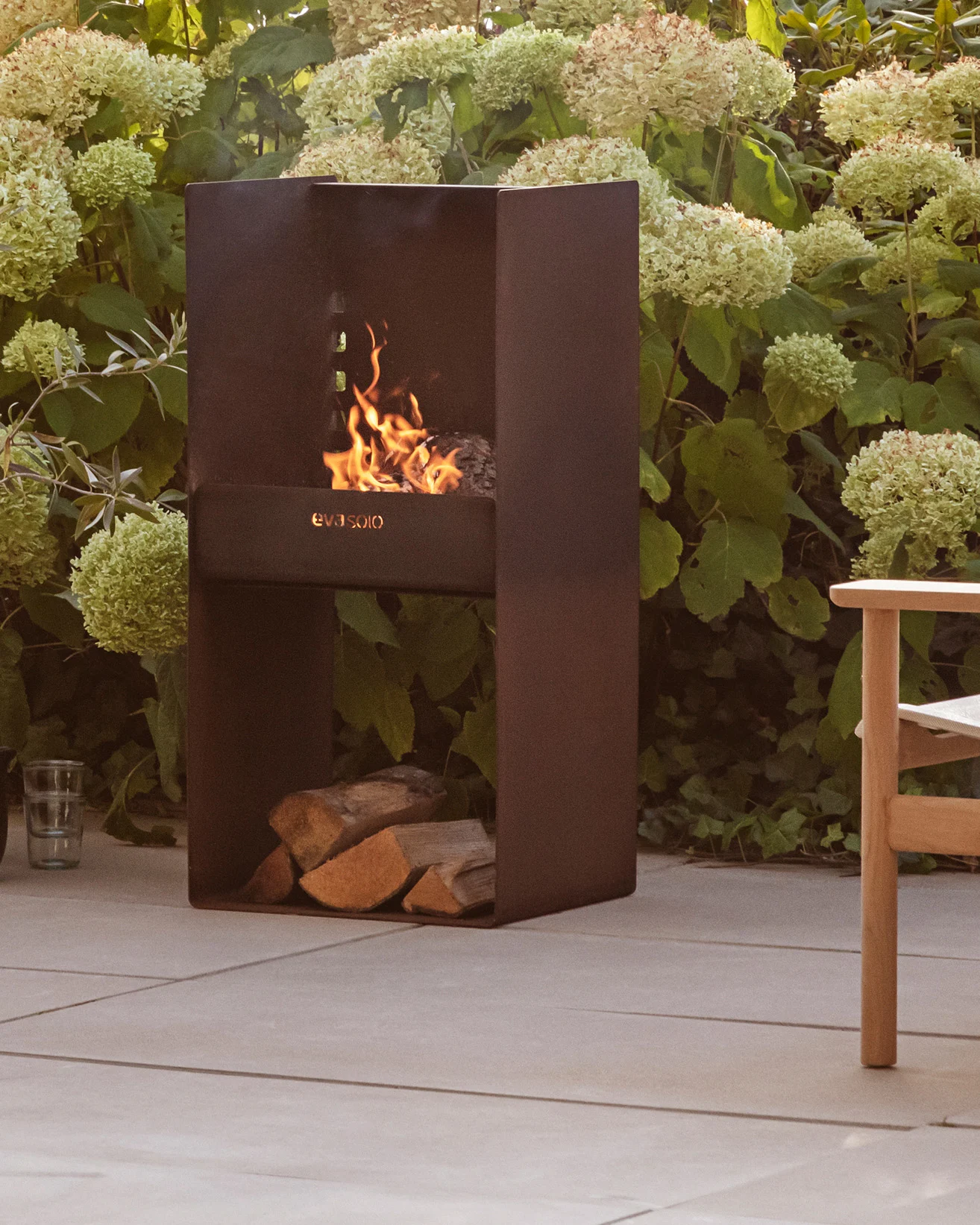 Moderni tumma metallinen ulkotakka, jossa palaa tuli, alla pinottuja puita, vihreiden kasvien ja terassin ympäröimänä.