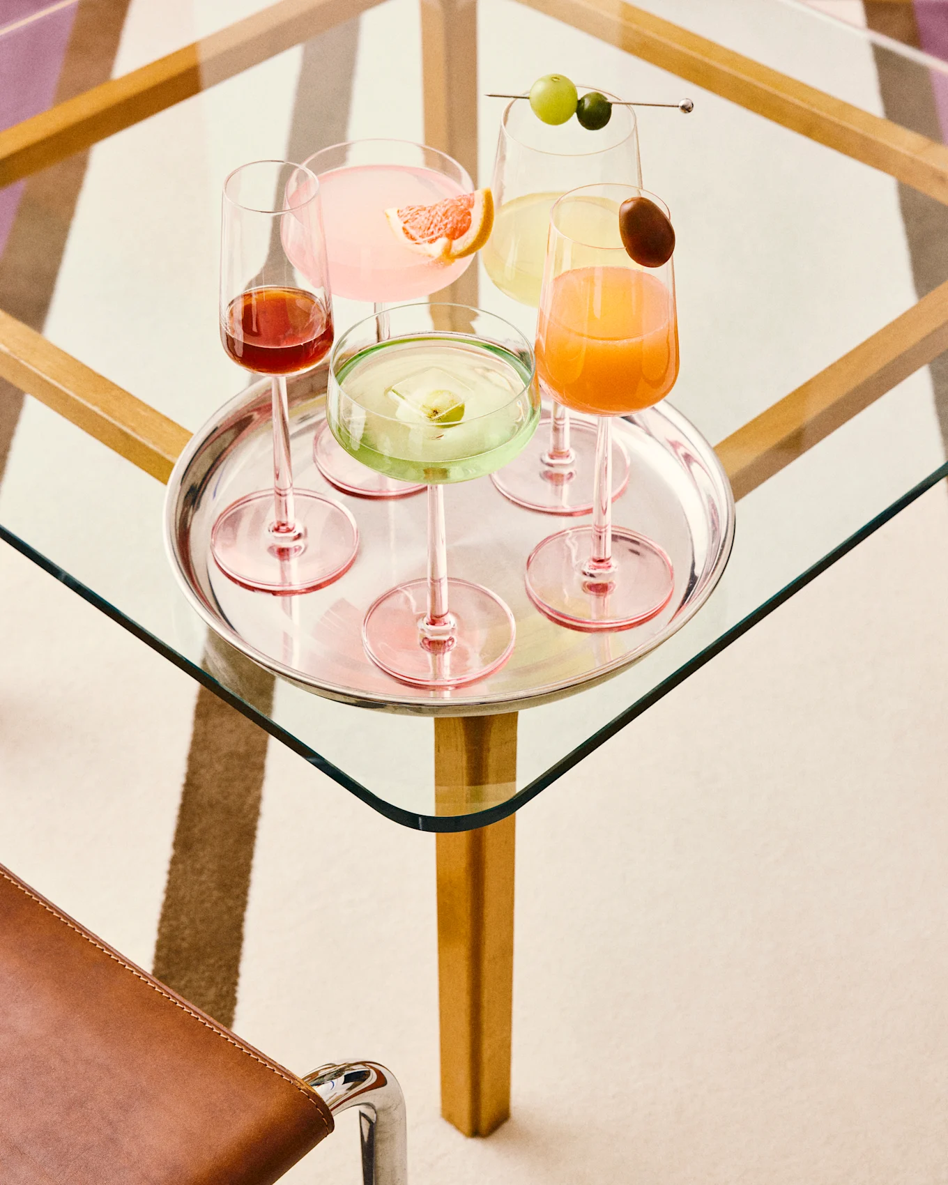 Viisi värikästä cocktailia hopeatarjottimella: pinkki greipin kera, vihreä hedelmäjään kera, keltainen viinirypäleiden kera, oranssi koristeella ja tummanpunainen, kaikki lasipöydällä.