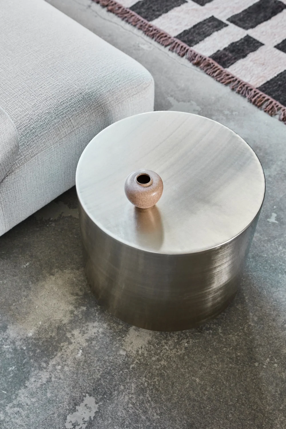 Pyöreä metallinen sivupöytä ja keraaminen maljakko, vaaleanharmaan sohvan ja kuvioidun maton vieressä betonilattialla.