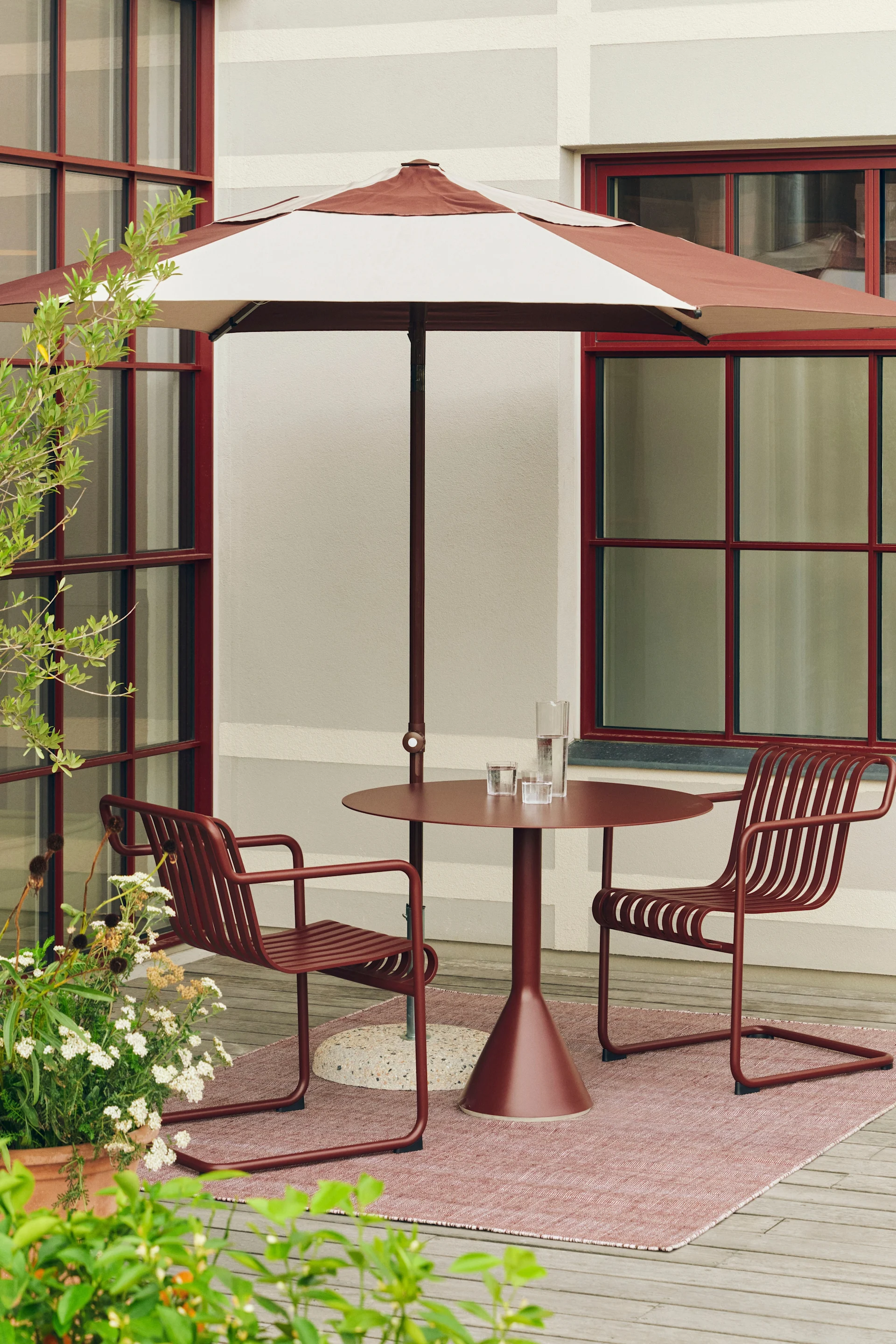 Ulkotarassi, jossa punaruskea raidallinen aurinkovarjo, pyöreä pöytä ja kaksi rimoitettua tuolia vaaleanpunaisella matolla.