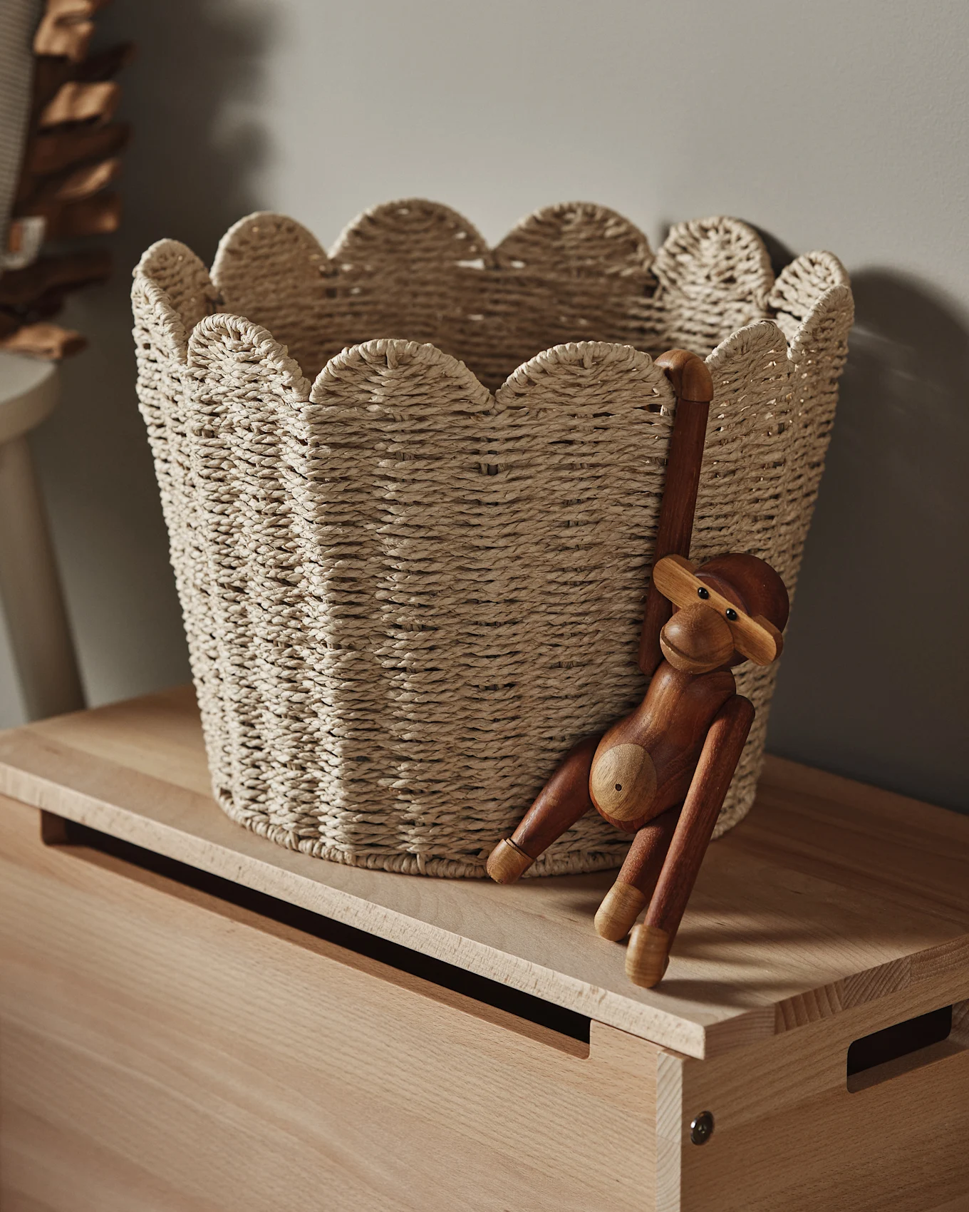 Vaalea punottu kori, jossa on aaltoileva reuna, puupinnalla, puinen apinahahmo roikkuu sen sivulta.