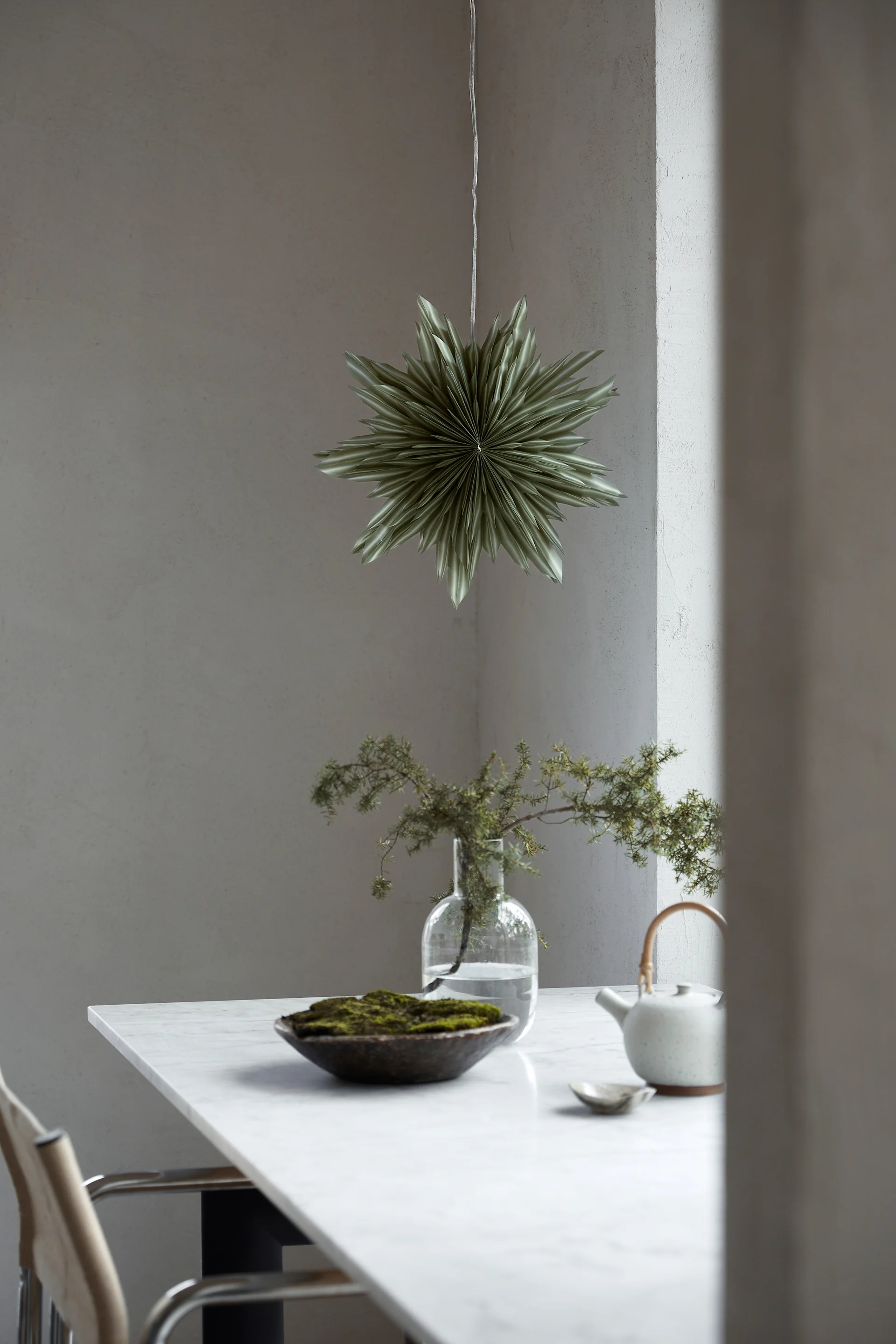 Vihreä paperinen tähtilyhty Helena roikkuu ikkunassa vihreän ruukkukasvin yläpuolella. 