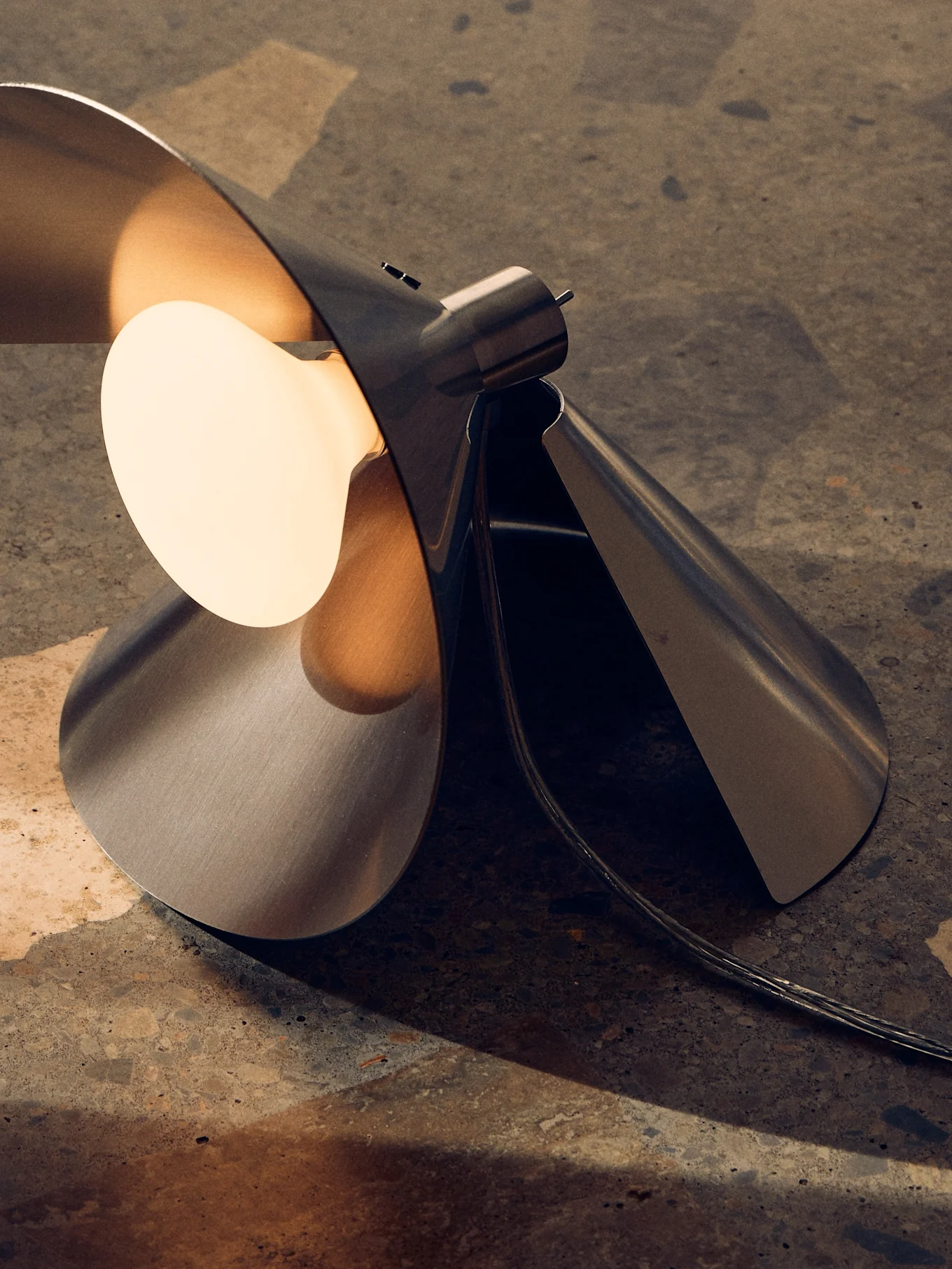 Moderni harjattu metallinen pöytävalaisin, veistoksellinen taitettu design ja lämmin, avonainen lamppu tekstuuripinnalla.
