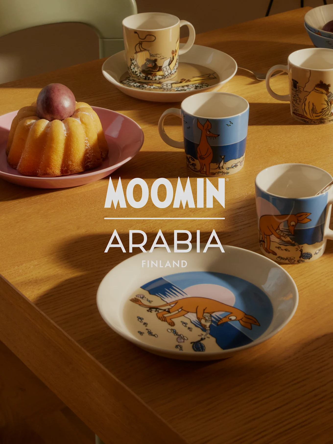 Moomin Arabia -astiasto puupöydällä kakun ja luumun kanssa. 'Moomin Arabia Finland' -logo.