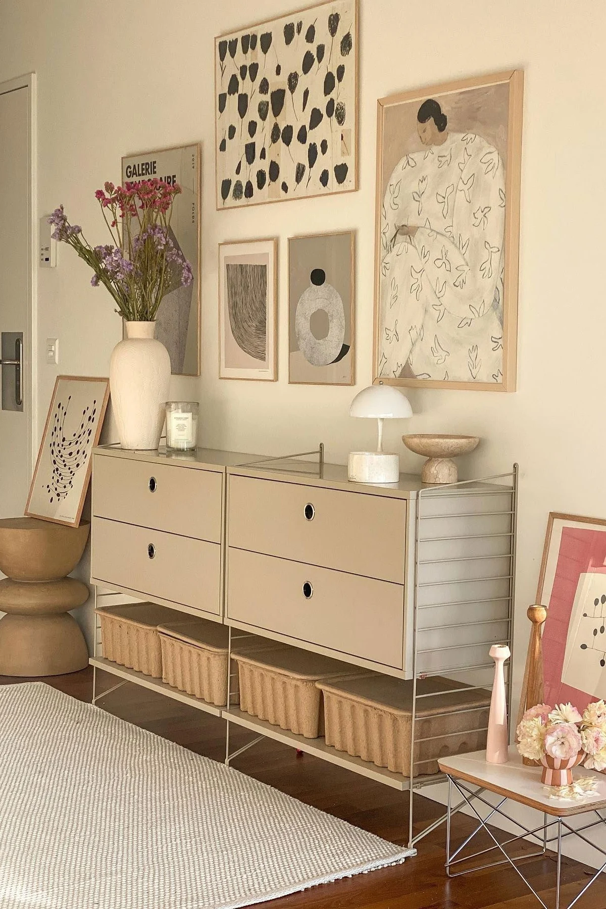 Moderni huone, jossa on beige modulaarinen kaappi, laatikot ja säilytyskorit. Maljakko kukkia, lamppu ja abstrakti taide koristavat tilaa.