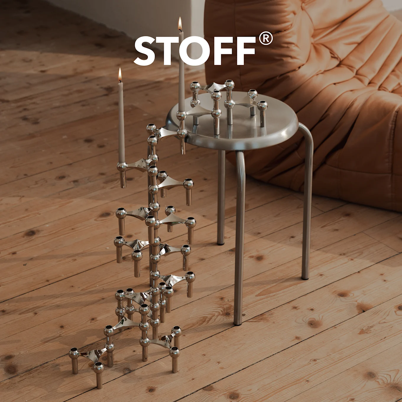 STOFF-logo kromisen kynttilänjalan yllä palavilla kynttilöillä puulattialla, toinen hopeisella jakkaralla.