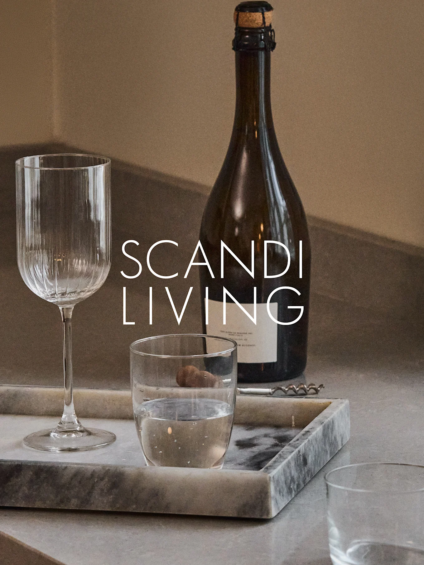 Tumma pullo, uurrettu viinilasi, vesilasi ja korkkiruuvi marmoritarjottimella, jossa on teksti 'SCANDI LIVING'.