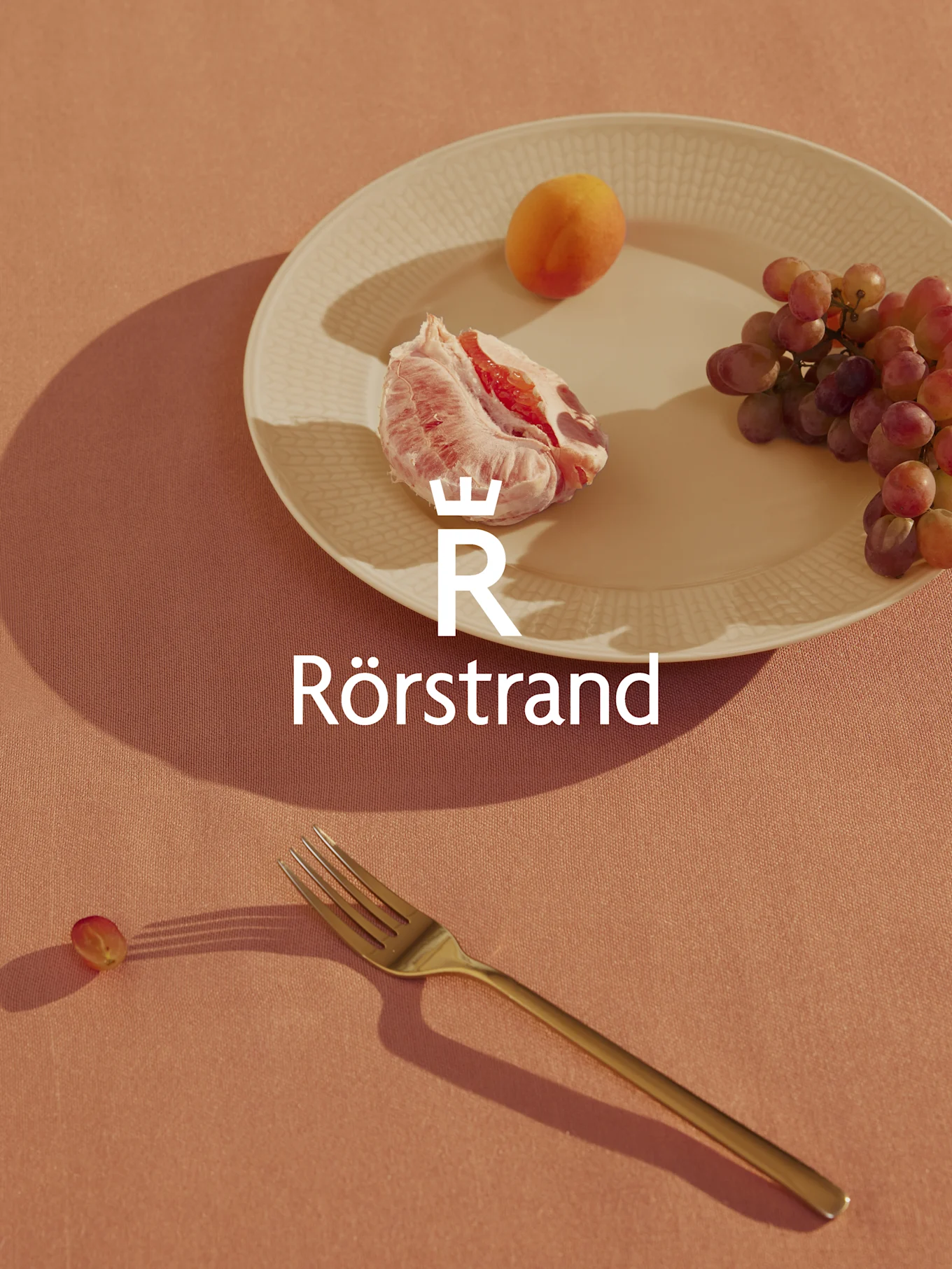 Rörstrand-logo lautasen päällä, jossa on greippiä, aprikoosia ja viinirypäleitä, haarukka ja yksi rypäle persikanvärisellä pöytäliinalla.