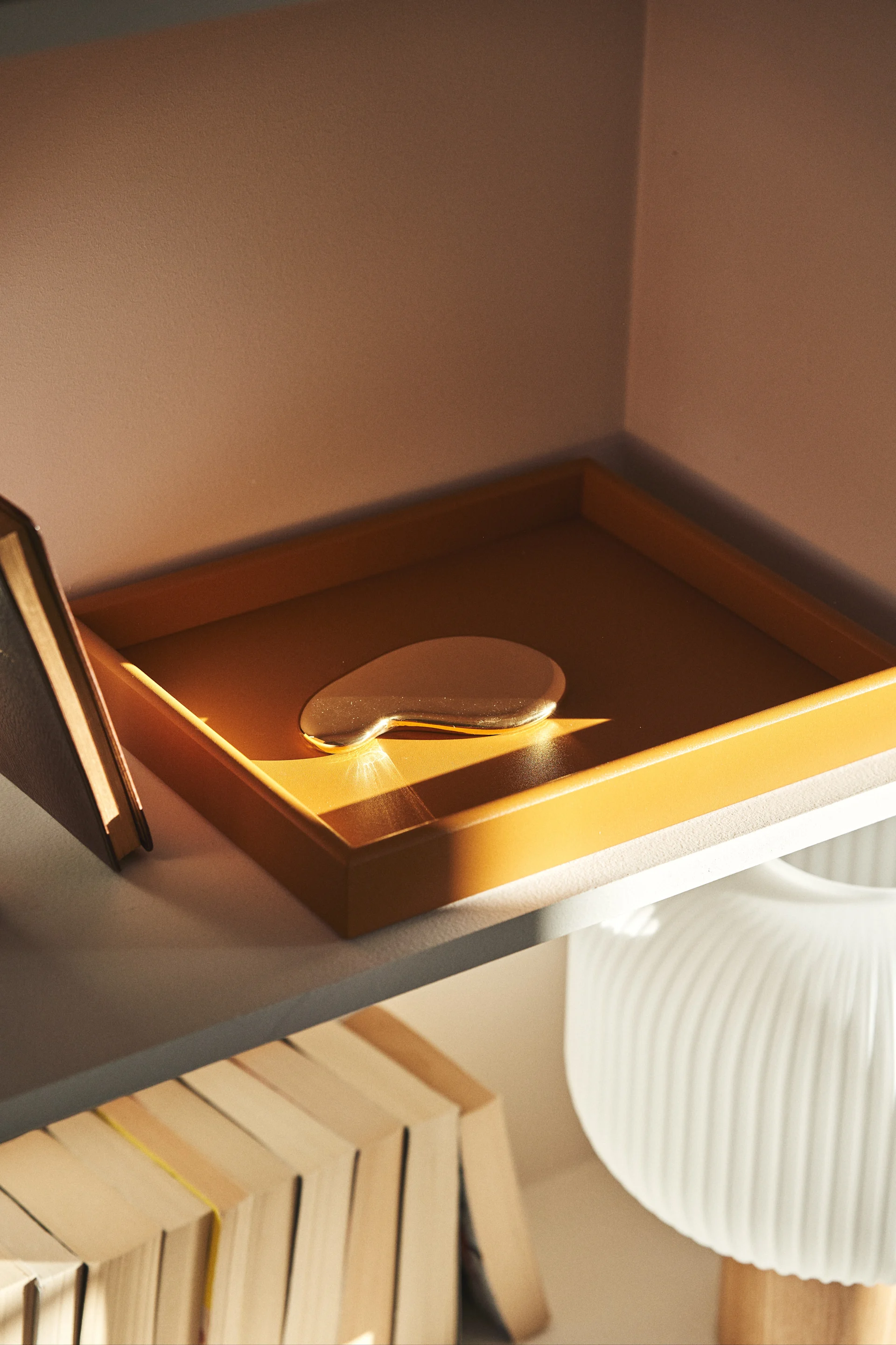 Tumma sinapinvärinen tarjotin seisoo puuhyllyllä auringonpaisteessa. Tarjotin on ihanteellinen tapa järjestää olohuone. 