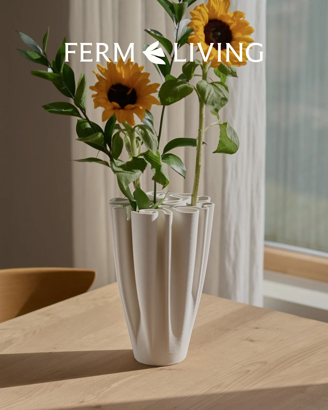Auringonkukkia valkoisessa, uurretussa aaltomaljakossa puupöydällä. Ferm Living -logo.